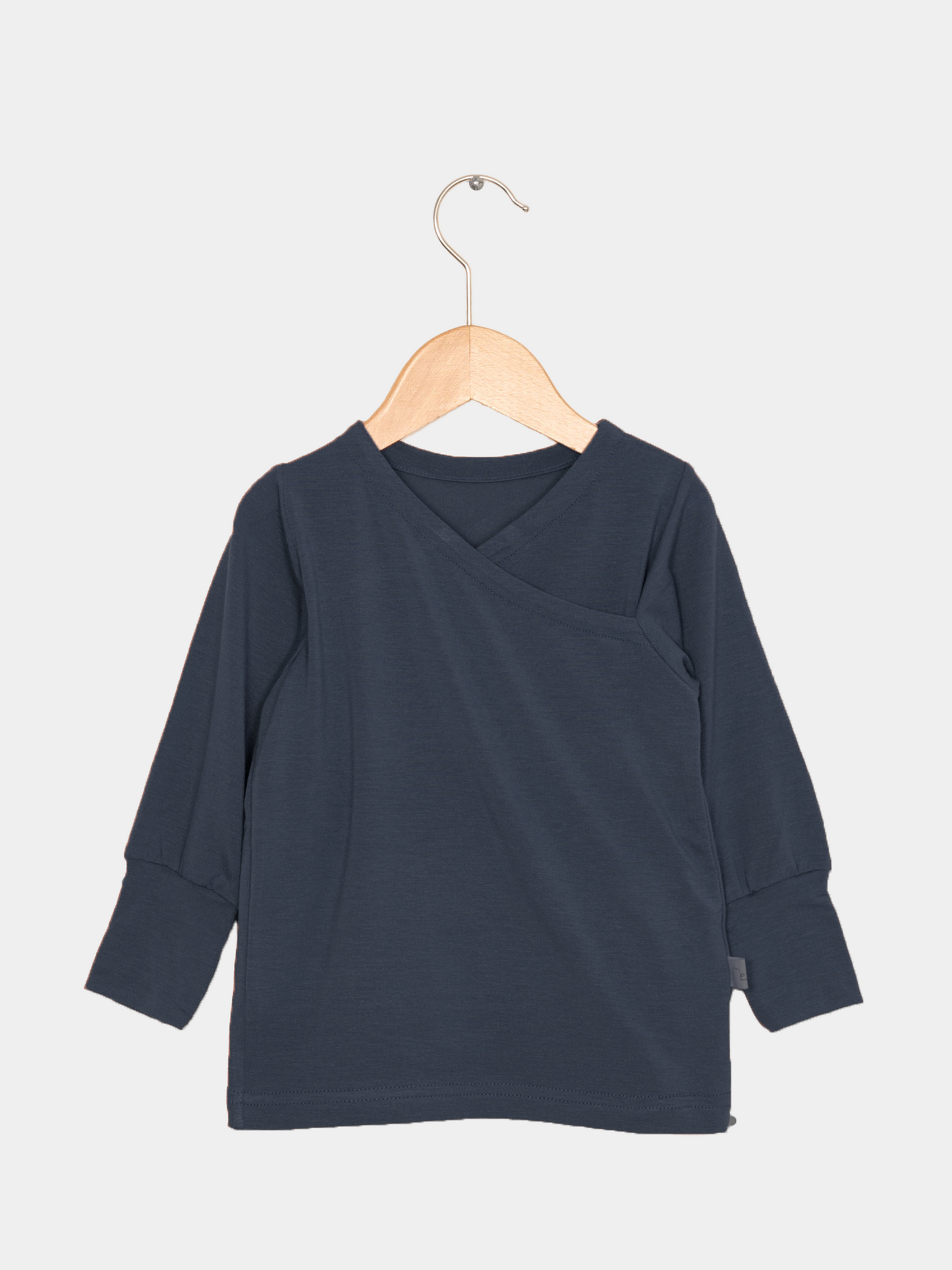 Wickelshirt aus Kaschmir-Mix - Blaubeere: Ein langärmeliges, dunkelblaues Shirt mit kimonoartigem Ausschnitt und gerippten Bündchen, hängend auf einem Holzkleiderbügel vor hellem Hintergrund.