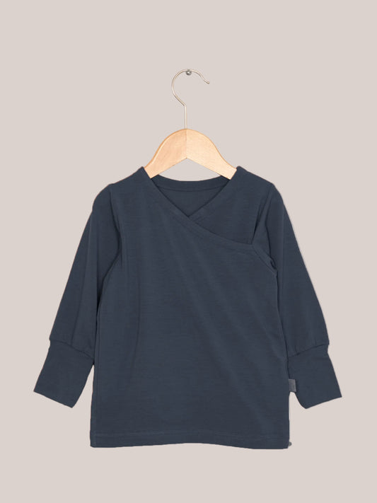 Wickelshirt aus Kaschmir-Mix - Blaubeere: Ein langärmeliges, dunkelblaues Shirt mit kimonoartigem Ausschnitt und gerippten Bündchen, hängend auf einem Holzkleiderbügel vor hellem Hintergrund.