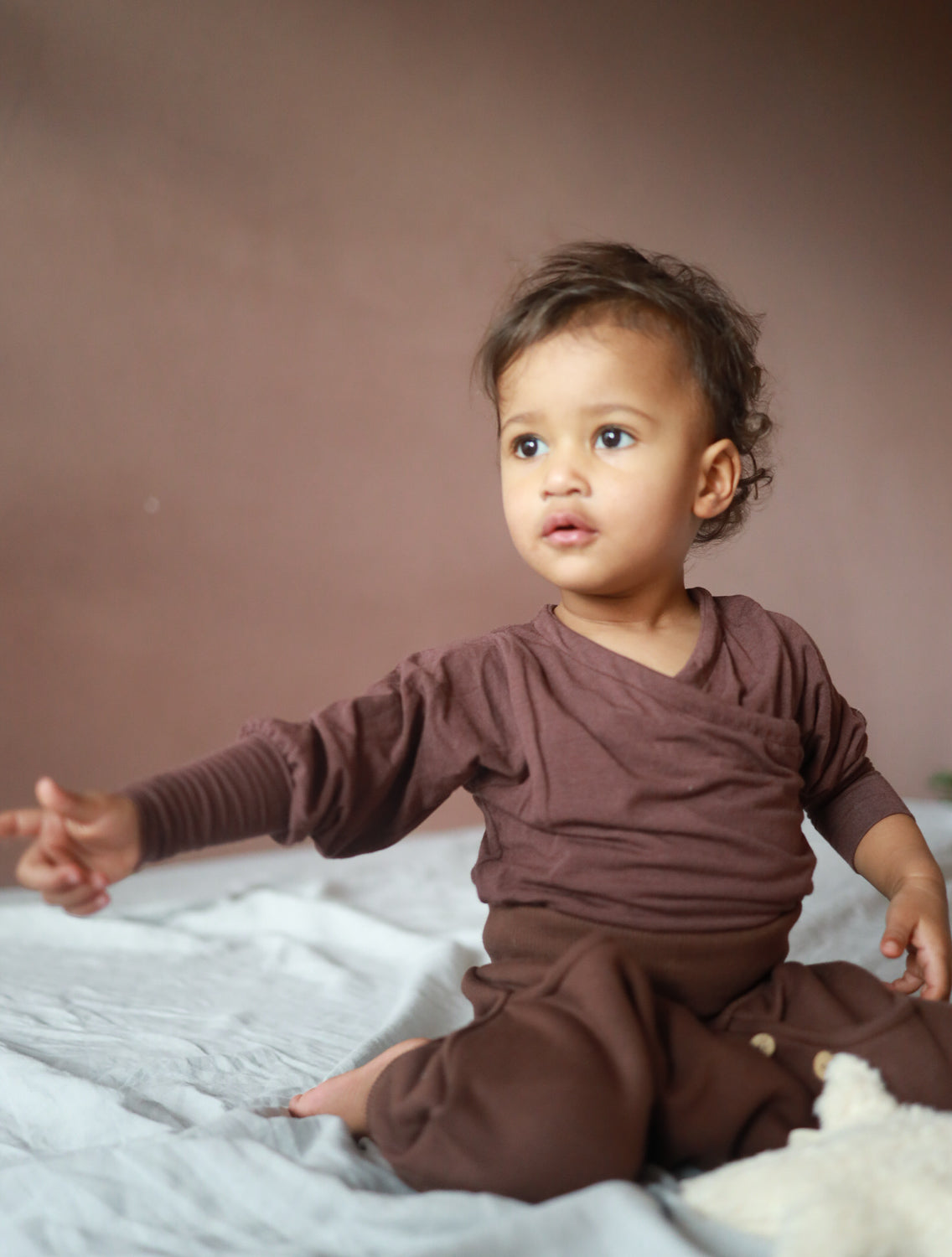 Ein Kleinkind sitzt auf einem Bett und zeigt auf etwas, während es das Wickelshirt aus Kaschmir-Mix - Malve trägt, das weich und dehnbar ist, ideal für Komfort und Mitwachsen. Kinderkleidung made in Berlin