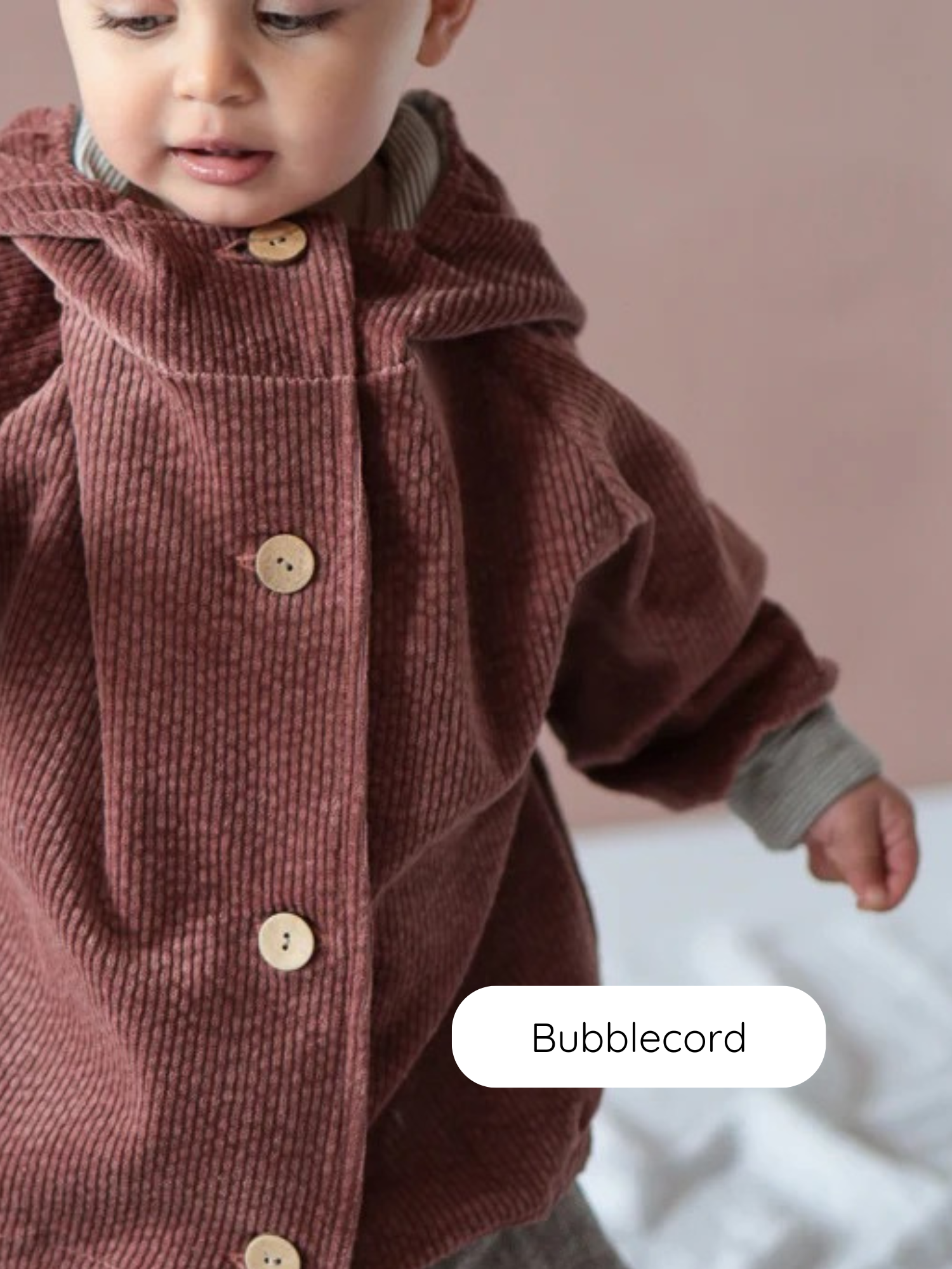 Kleinkind trägt die Wichteljacke Breitcord Stretch-Rosenholz mit süßer Wichtelkapuze. Die Jacke hat krempelbare Ringel-Armbündchen und wird mit hellen Holzknöpfen geschlossen.
