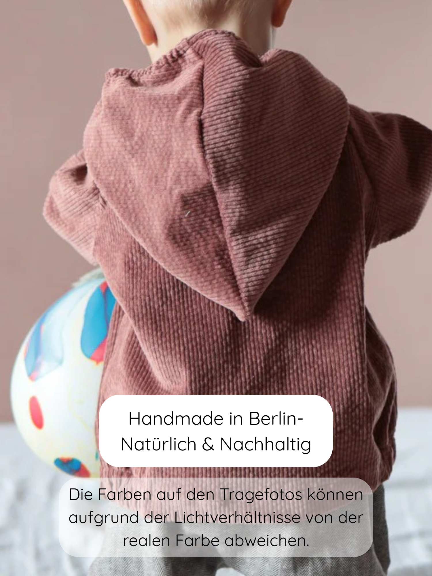 Ein Kleinkind trägt die Wichteljacke Breitcord Stretch-Rosenholz, eine braune Cordjacke mit Kapuze, und lehnt an einem farbenfrohen Ball. Die Jacke ist weich und hat Ringel-Armbündchen.