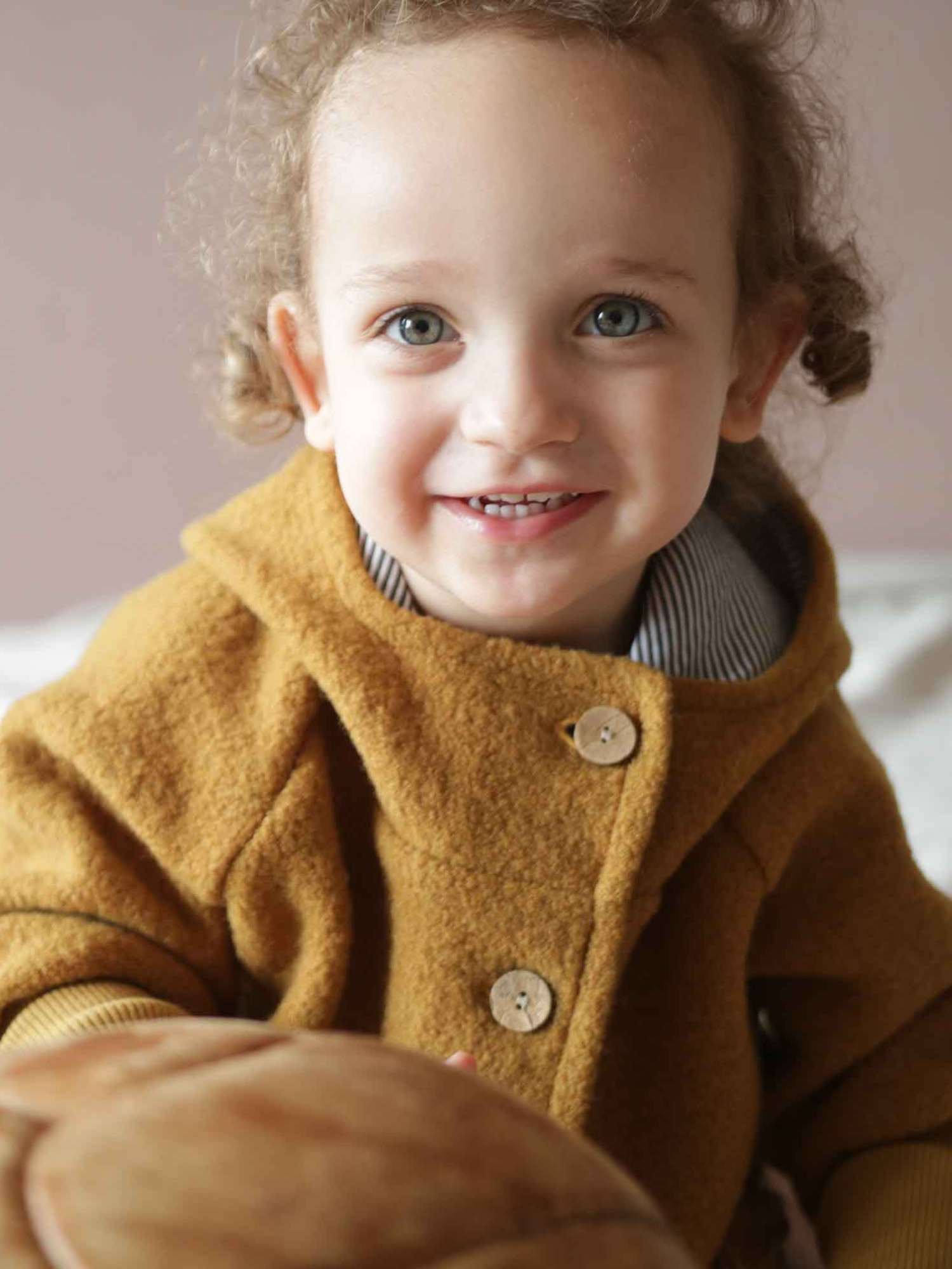 Ein lächelndes Kleinkind trägt die Wichteljacke Wollwalk - Honig mit Holzknöpfen und hält einen weichen Gegenstand. Die Jacke bietet Wärme und Bewegungsfreiheit, ideal für kühle Jahreszeiten.