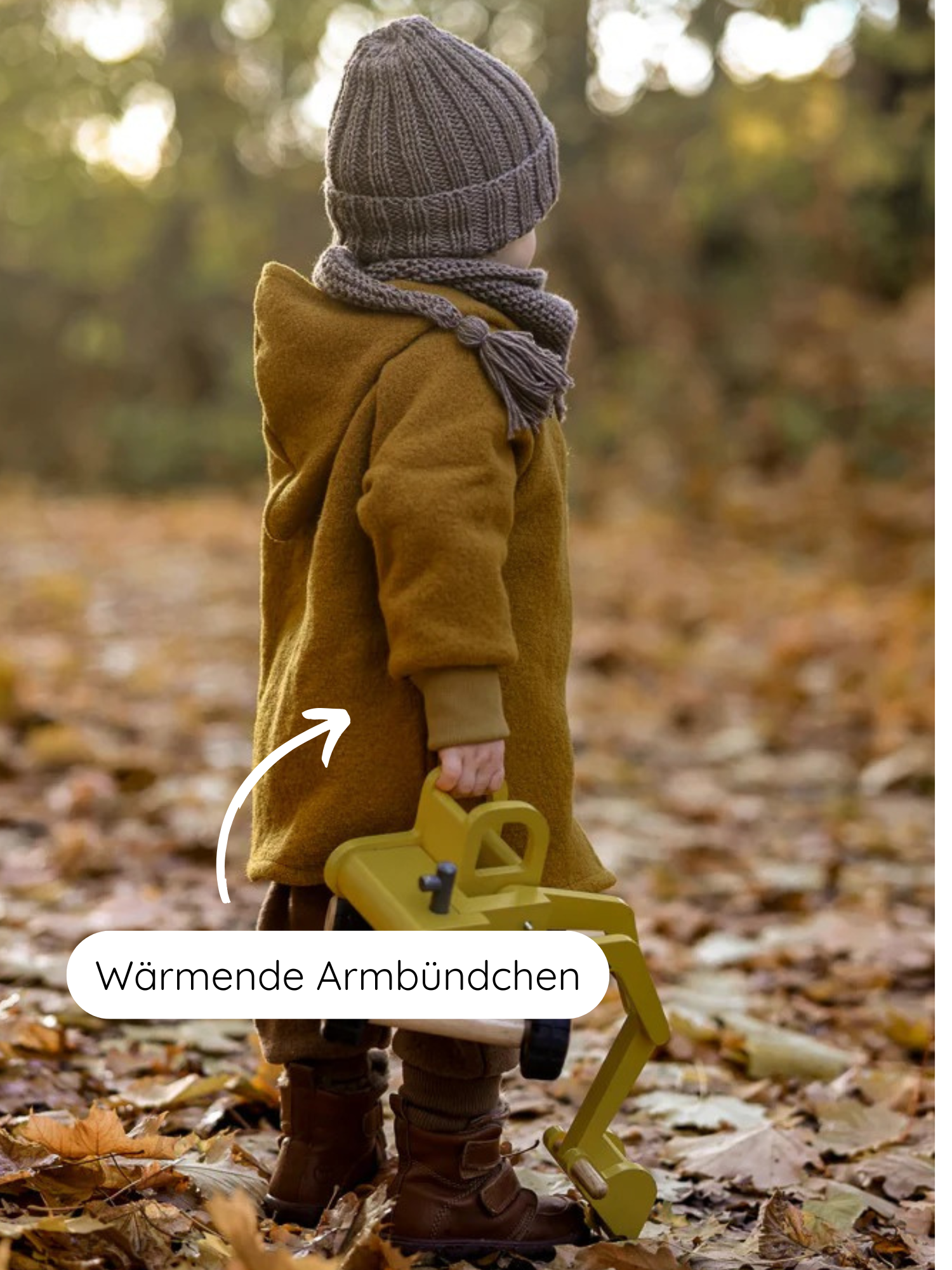Kind trägt die Wichteljacke Wollwalk - Honig mit wärmenden Armbündchen. Dazu ein grauer Schal und Mütze, braune Hose und Stiefel, hält einen gelben Spielzeugbagger.