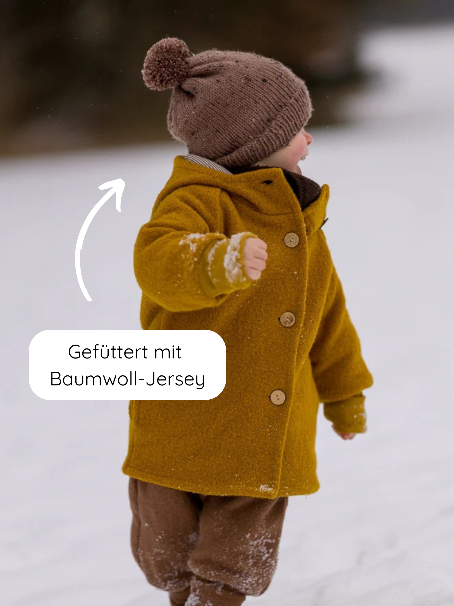 Kind im Schnee, trägt die Wichteljacke Wollwalk - Honig, eine senfgelbe Wolljacke mit Bio-Jersey-Futter, kombiniert mit braunem Strickmütze und Fleece-Hose. Perfekt für Herbst, Winter und Frühling.