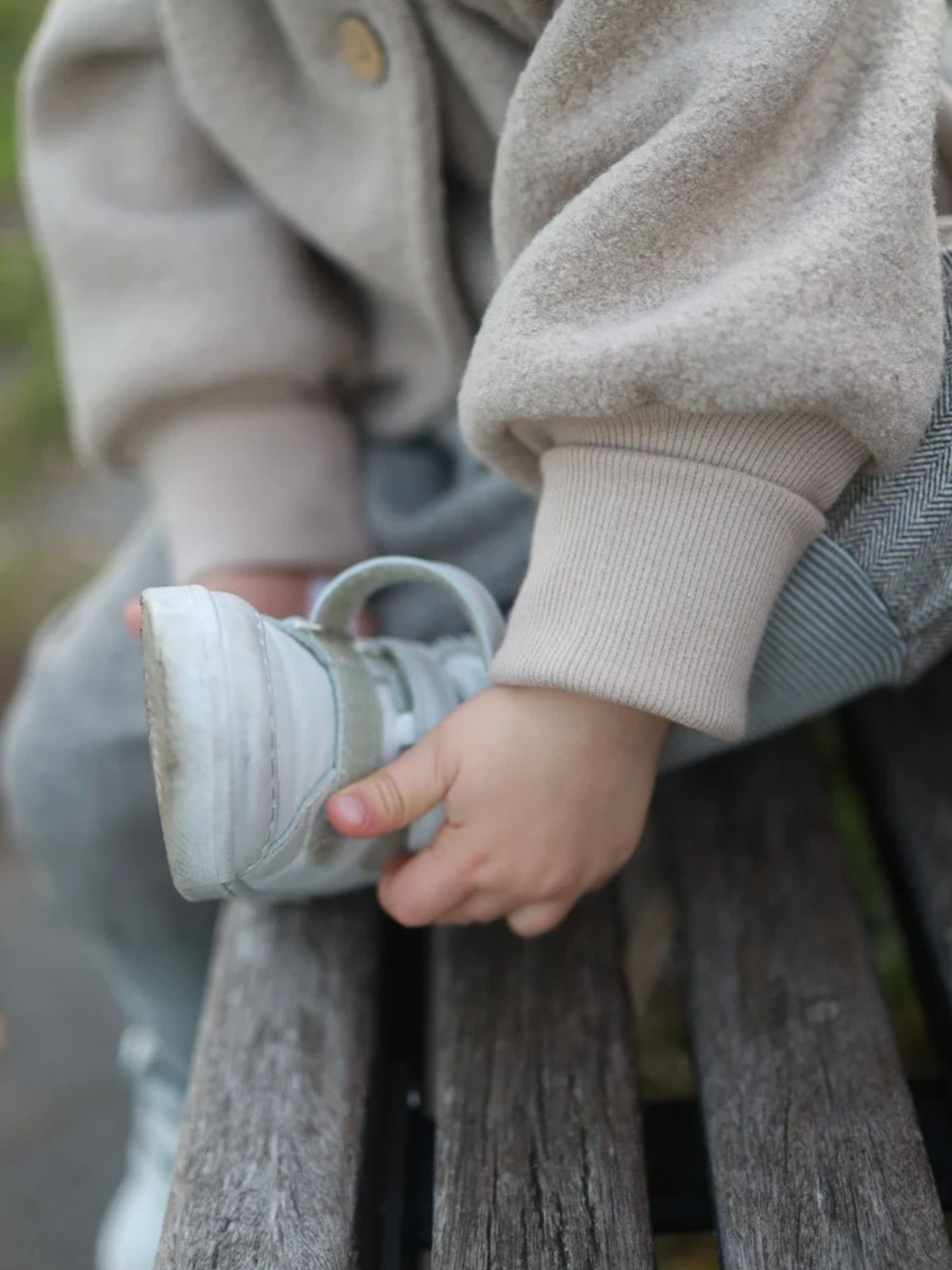 Ein Kleinkind sitzt auf einer Bank, trägt eine Wichteljacke Wollwalk - Sand mit krempelbaren Bündchen, während es an einem mintfarbenen Sneaker zieht. Die Jacke bietet Bewegungsfreiheit und Komfort.