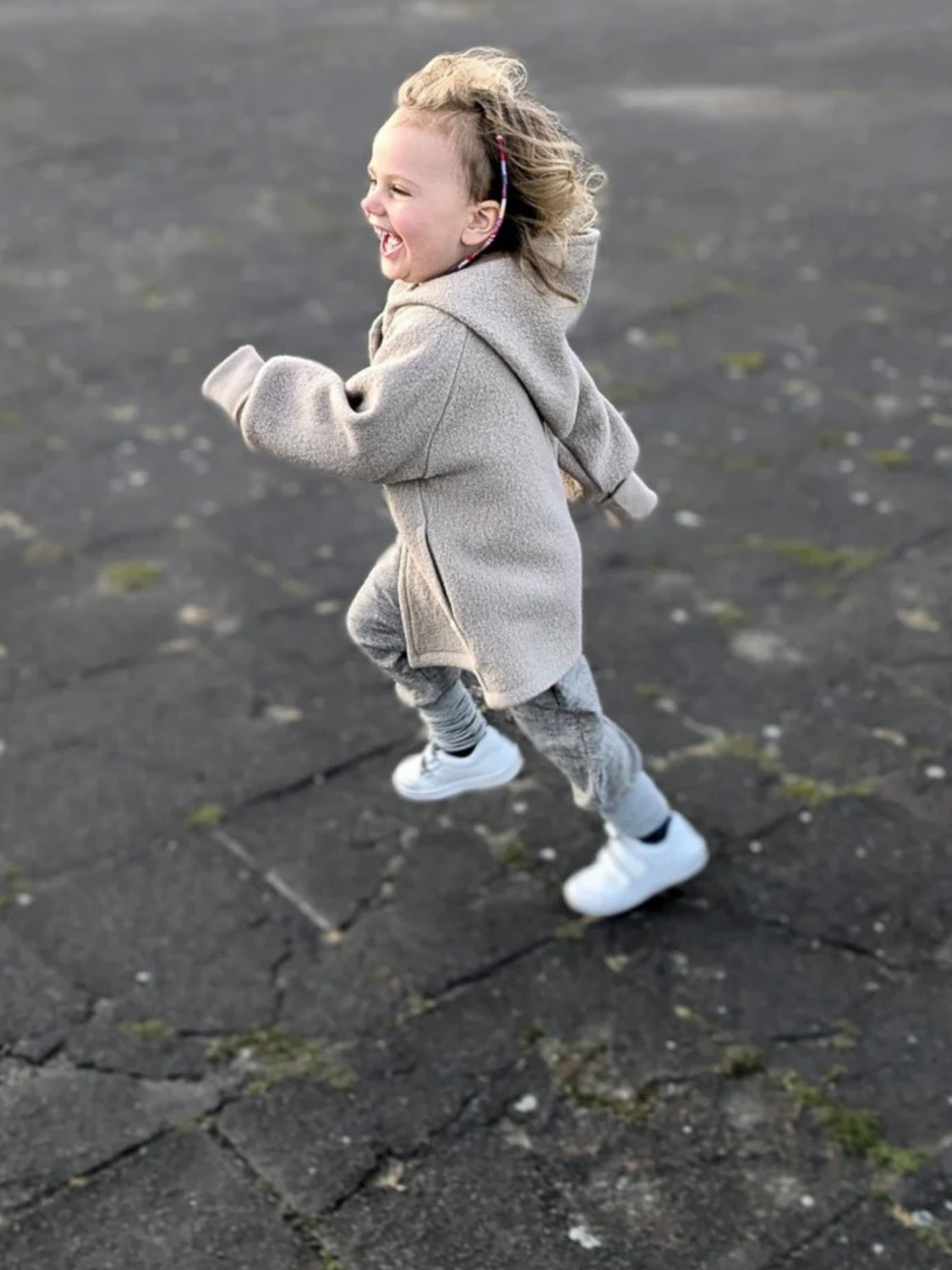 Ein Kleinkind rennt glücklich in der Wichteljacke Wollwalk - Sand. Die Jacke bietet Bewegungsfreiheit und ist gefüttert mit Bio-Jersey, ideal für Herbst, Frühling und Winter.