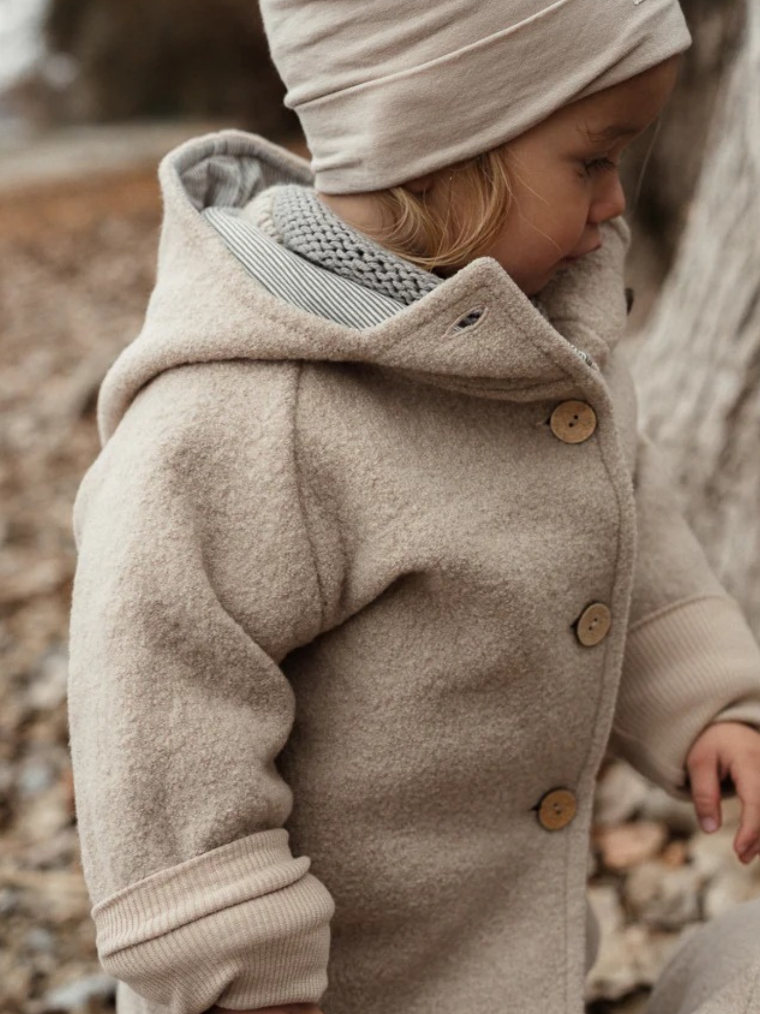 Ein Kind trägt die Wichteljacke Wollwalk - Sand mit Holzknöpfen, Kapuze und krempelbaren Bündchen. Die Jacke bietet Bewegungsfreiheit und ist ideal für kühle Jahreszeiten im Park.