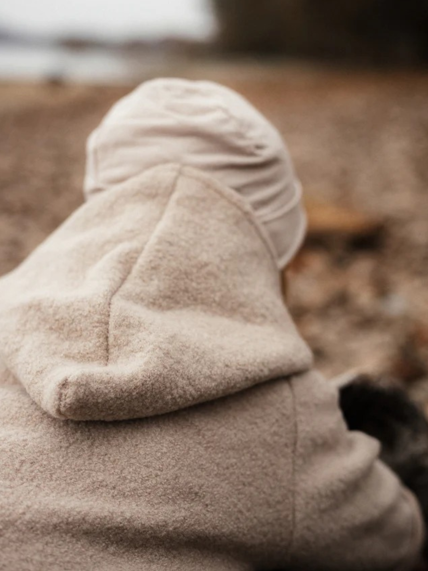 Ein Kleinkind trägt die Wichteljacke Wollwalk - Sand mit Kapuze, aus hochwertigem Wollwalk und Bio-Jersey-Futter, während es am Kiesstrand sitzt.