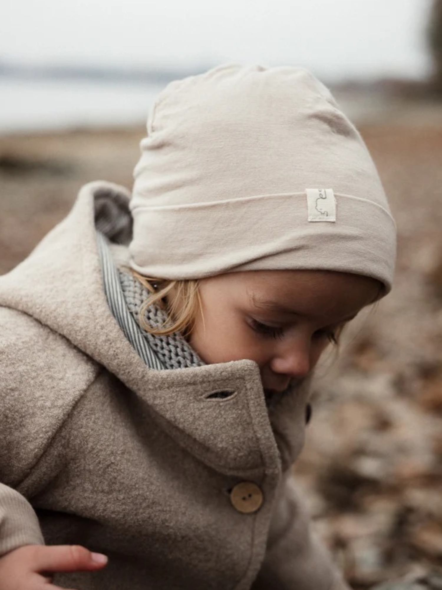 Ein Kind trägt die Wichteljacke Wollwalk - Sand mit Bio-Jersey-Futter, sichtbar sind der weiche beigefarbene Wollwalk-Stoff, ein Holzknopf und ein helles Beanie, ideal für Herbst und Winter.