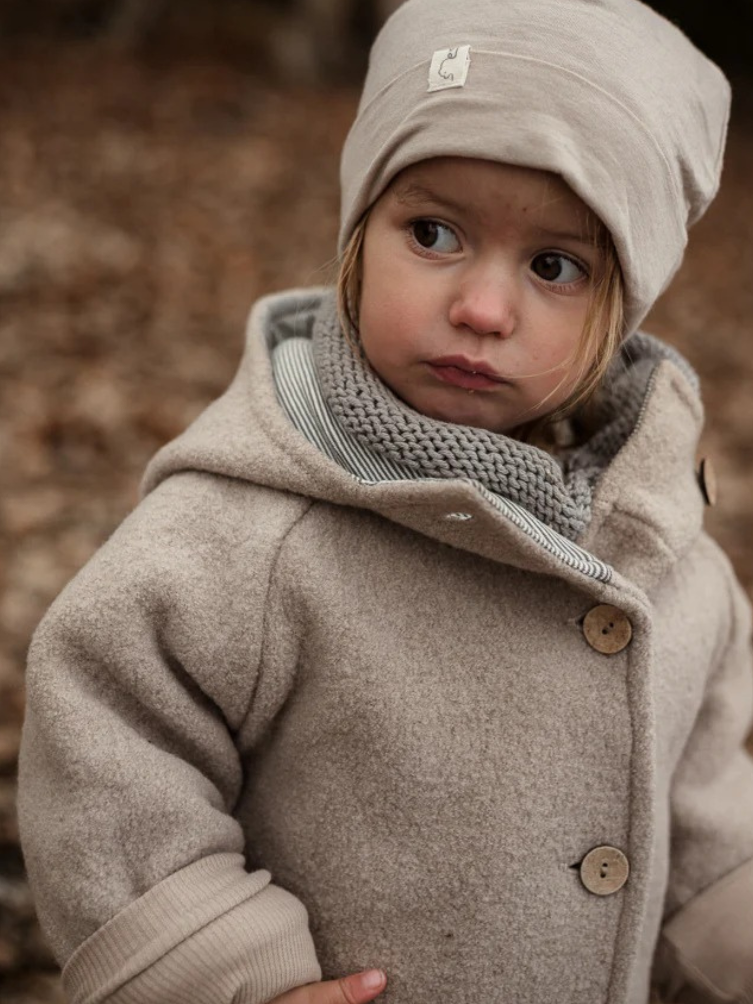 Kleinkind trägt die Wichteljacke Wollwalk - Sand mit großen Holzknöpfen, kombiniert mit passendem Strickschal und Beanie, steht nachdenklich im Freien auf Laubboden.