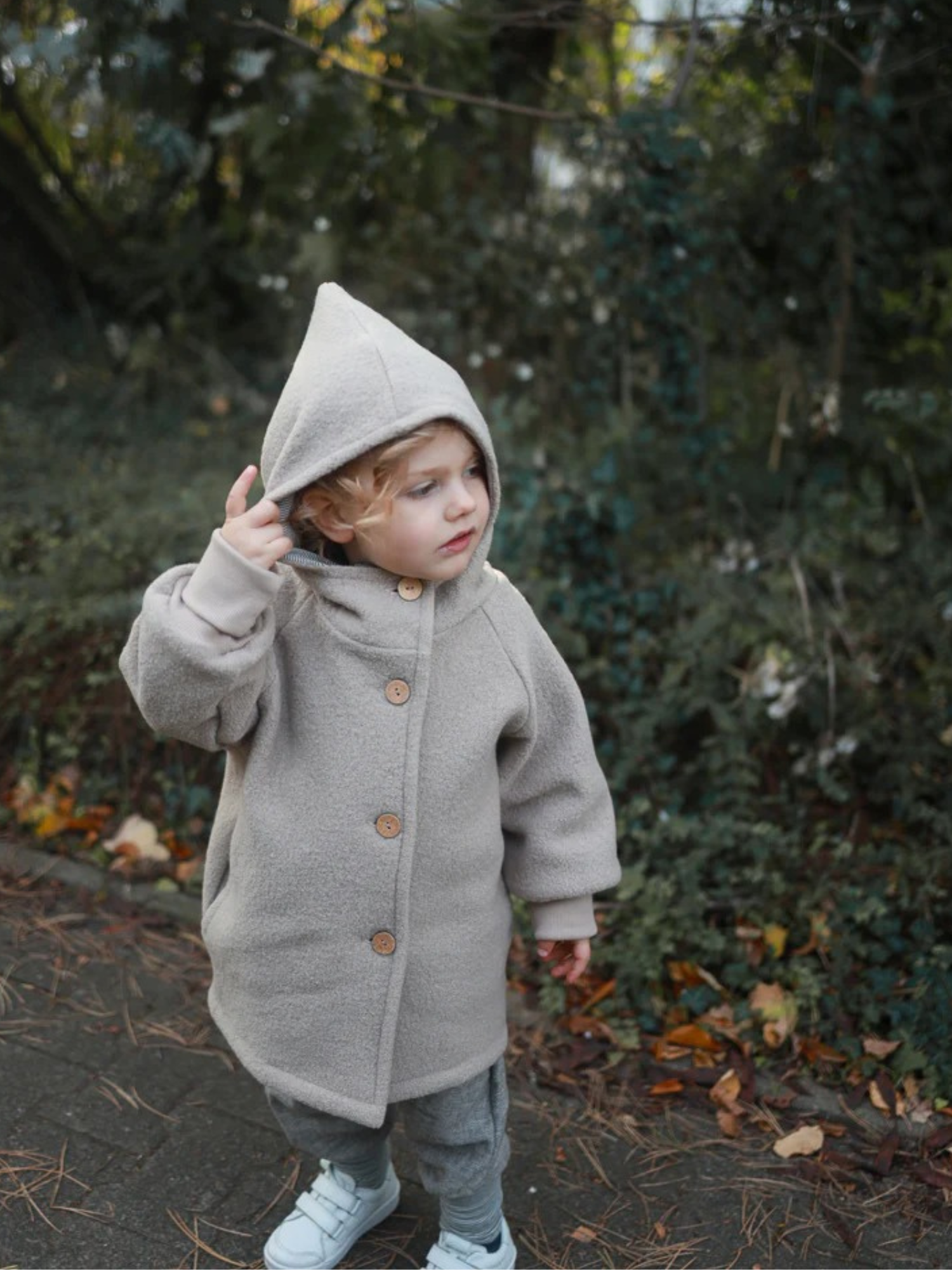 Kleinkind trägt die Wichteljacke Wollwalk - Sand, eine hellgrau-beige Wolljacke mit Holzknöpfen und Kapuze, auf einem herbstlichen Weg. Jacke aus Bio-Jersey gefüttert, bietet Bewegungsfreiheit und Temperaturregulierung.