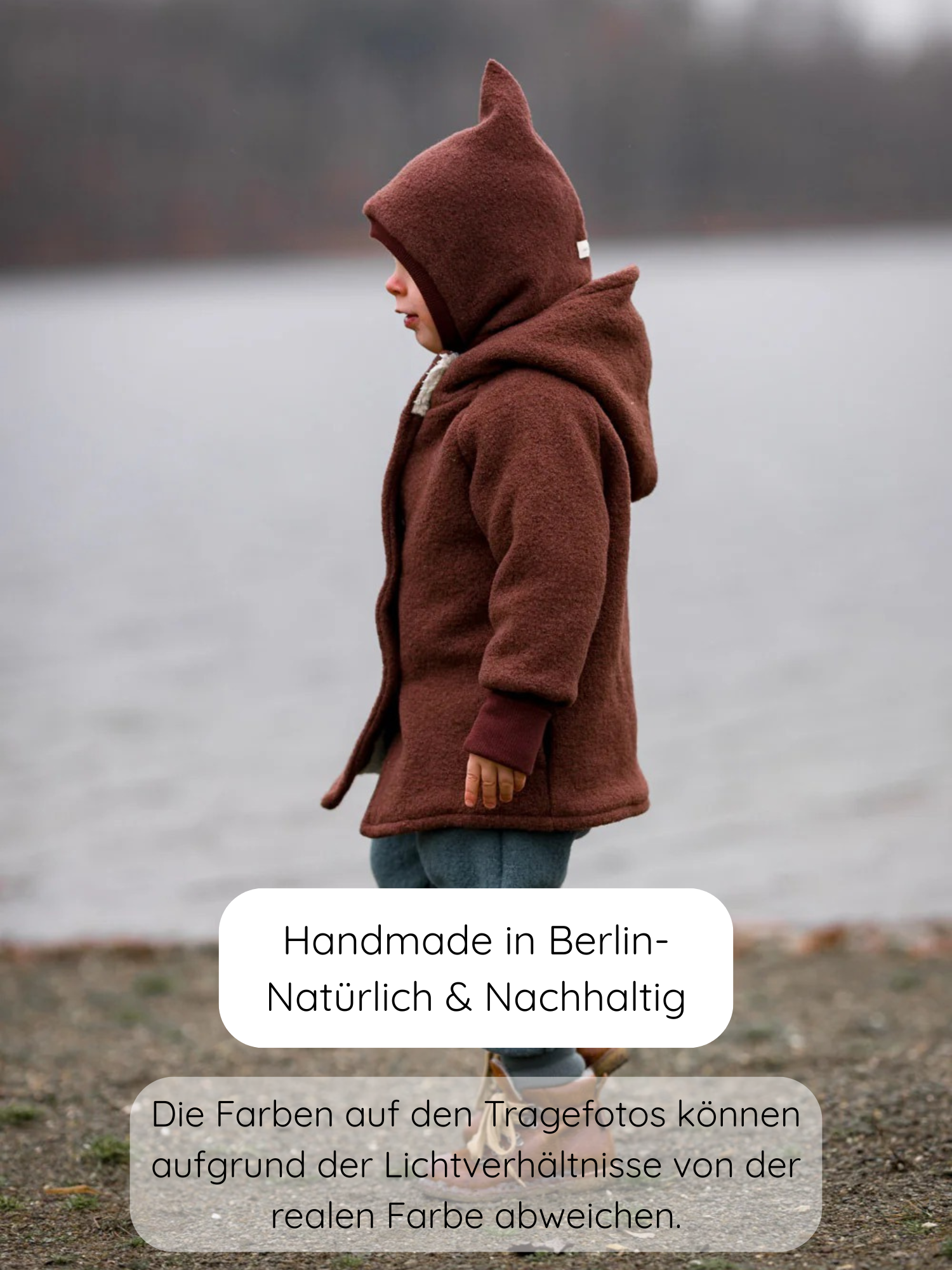 Kleinkind trägt die Wichteljacke Wollwalk - Schoko mit spitzen Kapuzenohren am steinigen Seeufer, präsentiert als handgefertigt und nachhaltig aus Berlin, ideal für Herbst bis Frühling.