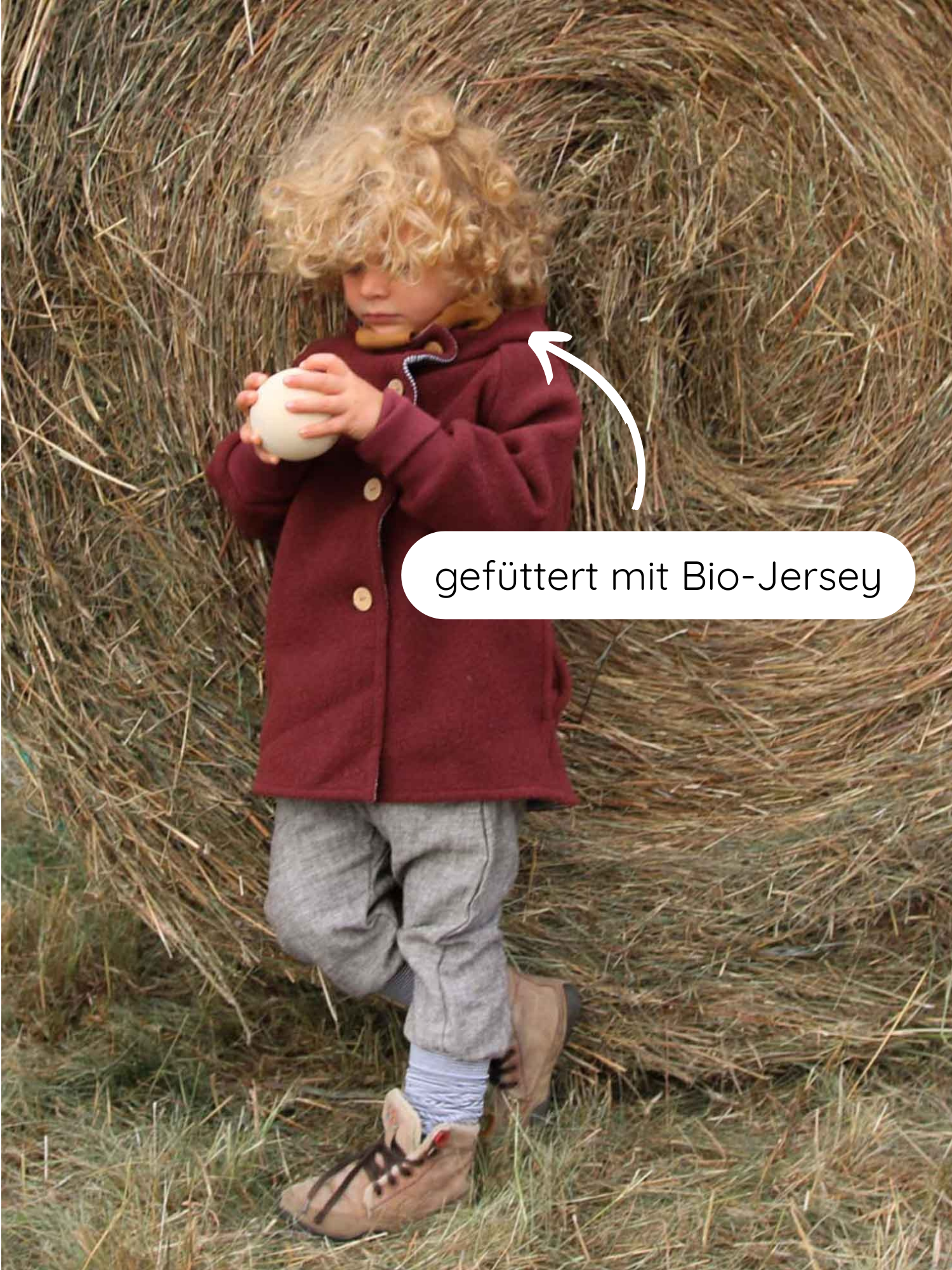 Ein Kind lehnt an einem Heuballen, trägt die Wichteljacke Wollwalk - Ziegel mit Kapuze, gefüttert mit Bio-Jersey. Die Jacke bietet Komfort und Bewegungsfreiheit.