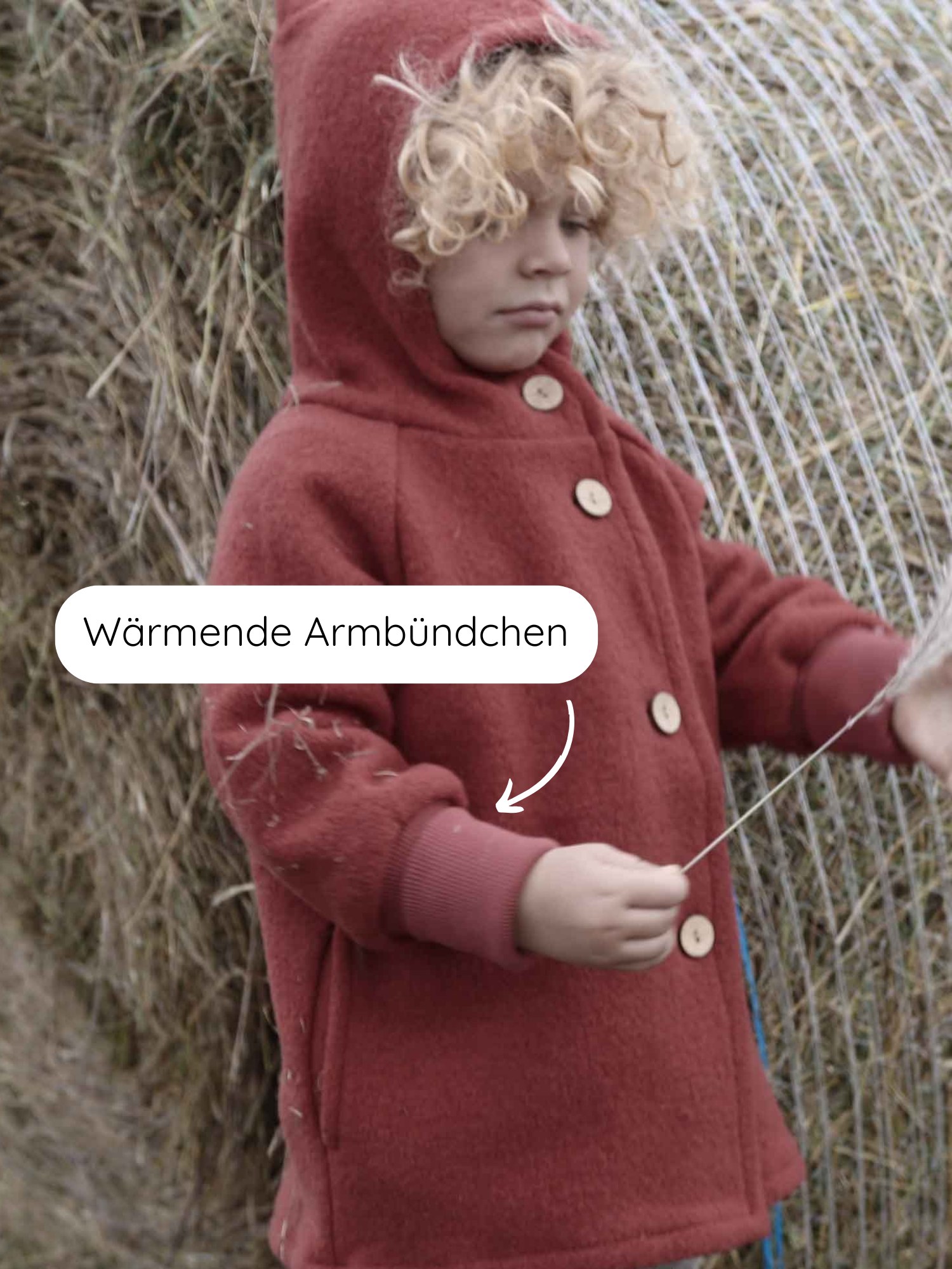 Ein Kind trägt die Wichteljacke Wollwalk - Ziegel, eine ziegelrote Wolljacke mit Kapuze, Holzknöpfen, und wärmenden Armbündchen, stehend vor einem großen Heuballen, hält einen Heustrang.