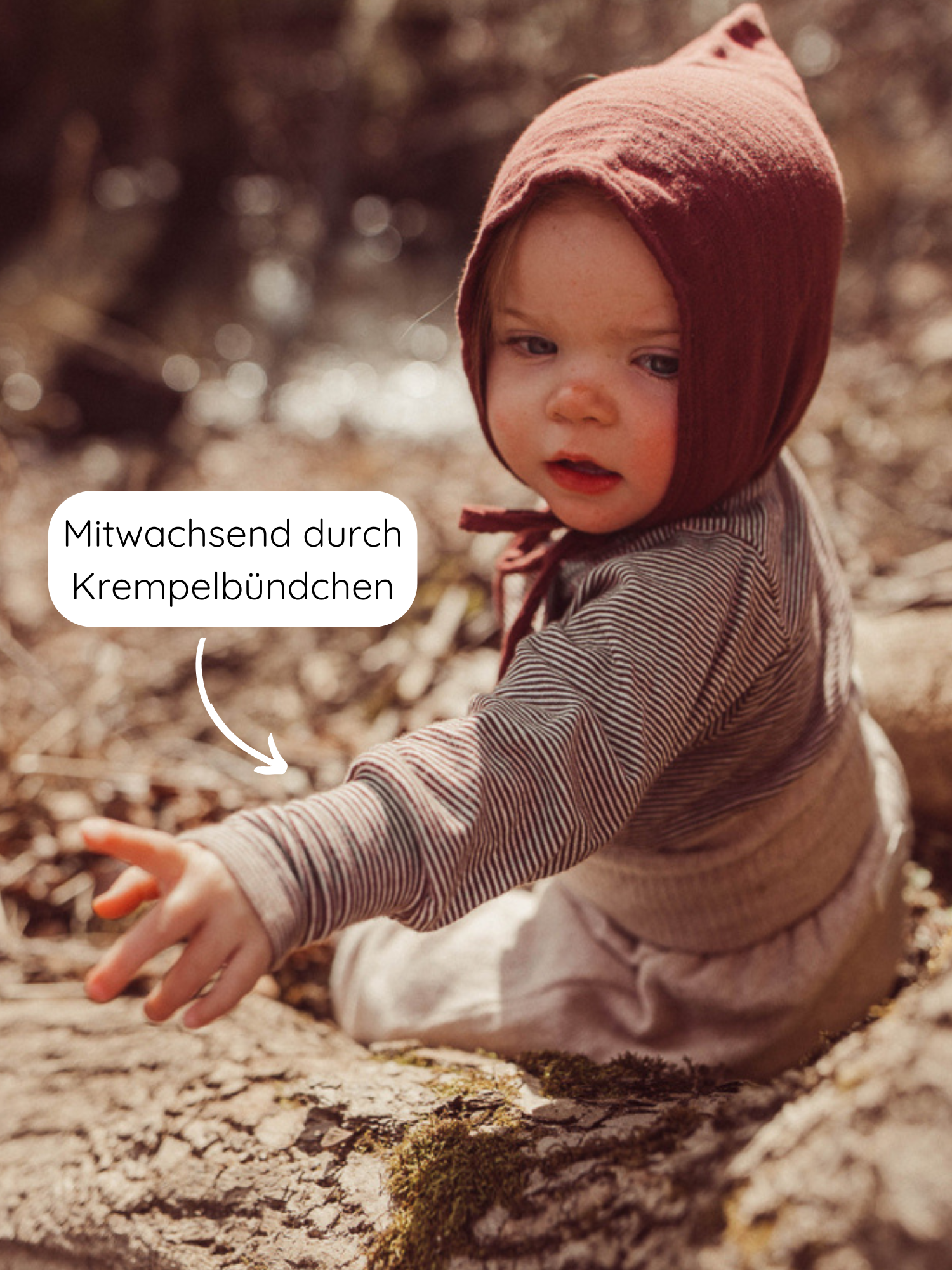 Kleinkind trägt ein Wickelshirt aus Bio-Baumwolle - Ringel Schoko mit krempelbaren Bündchen, auf einem Baumstamm sitzend. Pfeil zeigt auf die Ärmel mit dem Hinweis „Mitwachsend durch Krempelbündchen“.