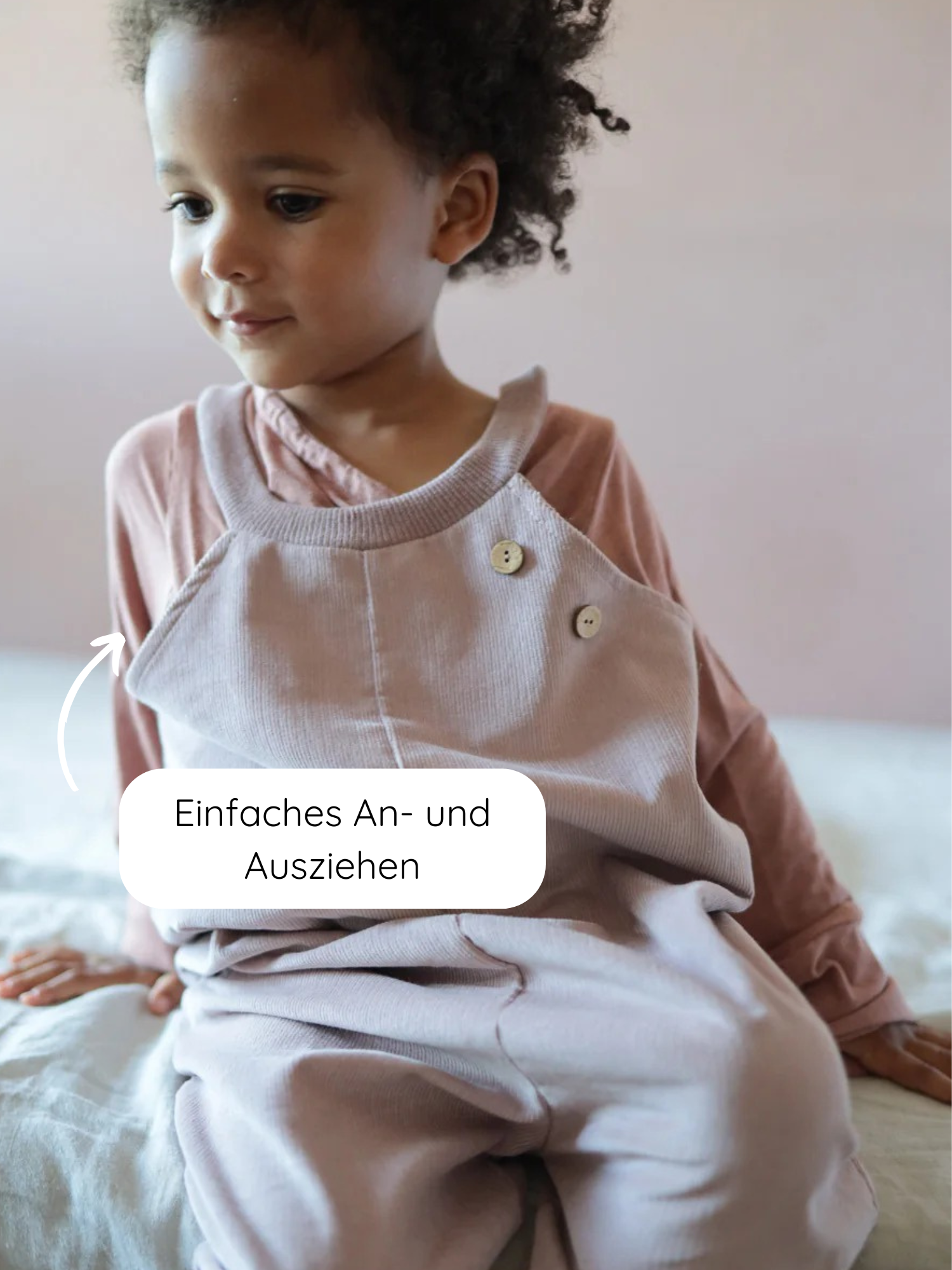 Kleinkind trägt das Wickelshirt aus Kaschmir-Mix - Zimt, kombiniert mit einem passenden Overall. Ein Hinweis zeigt auf einen Knopf, der das einfache An- und Ausziehen des Overalls hervorhebt.
