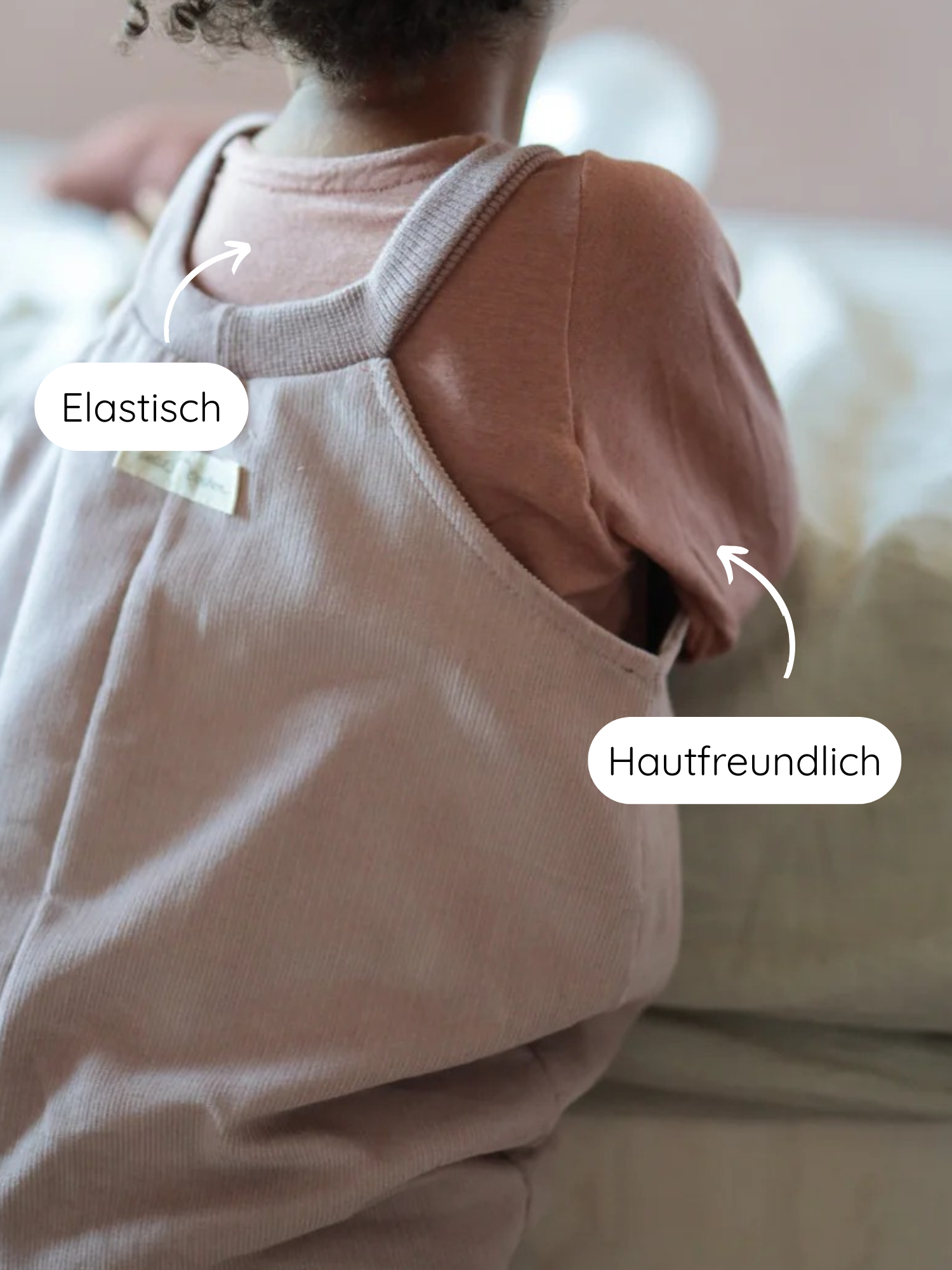 Kleines Kind im Wickelshirt aus Kaschmir-Mix - Zimt, sitzt auf weicher Oberfläche. Elastische Träger und hautfreundliche Armausschnitte betonen die weiche, mitwachsende Qualität des Materials.
