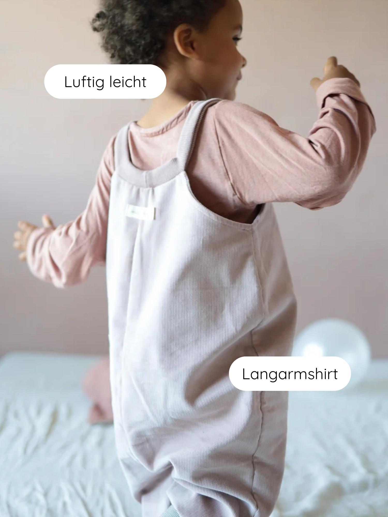 Kind trägt Wickelshirt aus Kaschmir-Mix - Zimt, über einem rosa Langarmshirt. Das Wickelshirt wirkt luftig-leicht, hinten im Bild unscharfes Bett und Ballon.
