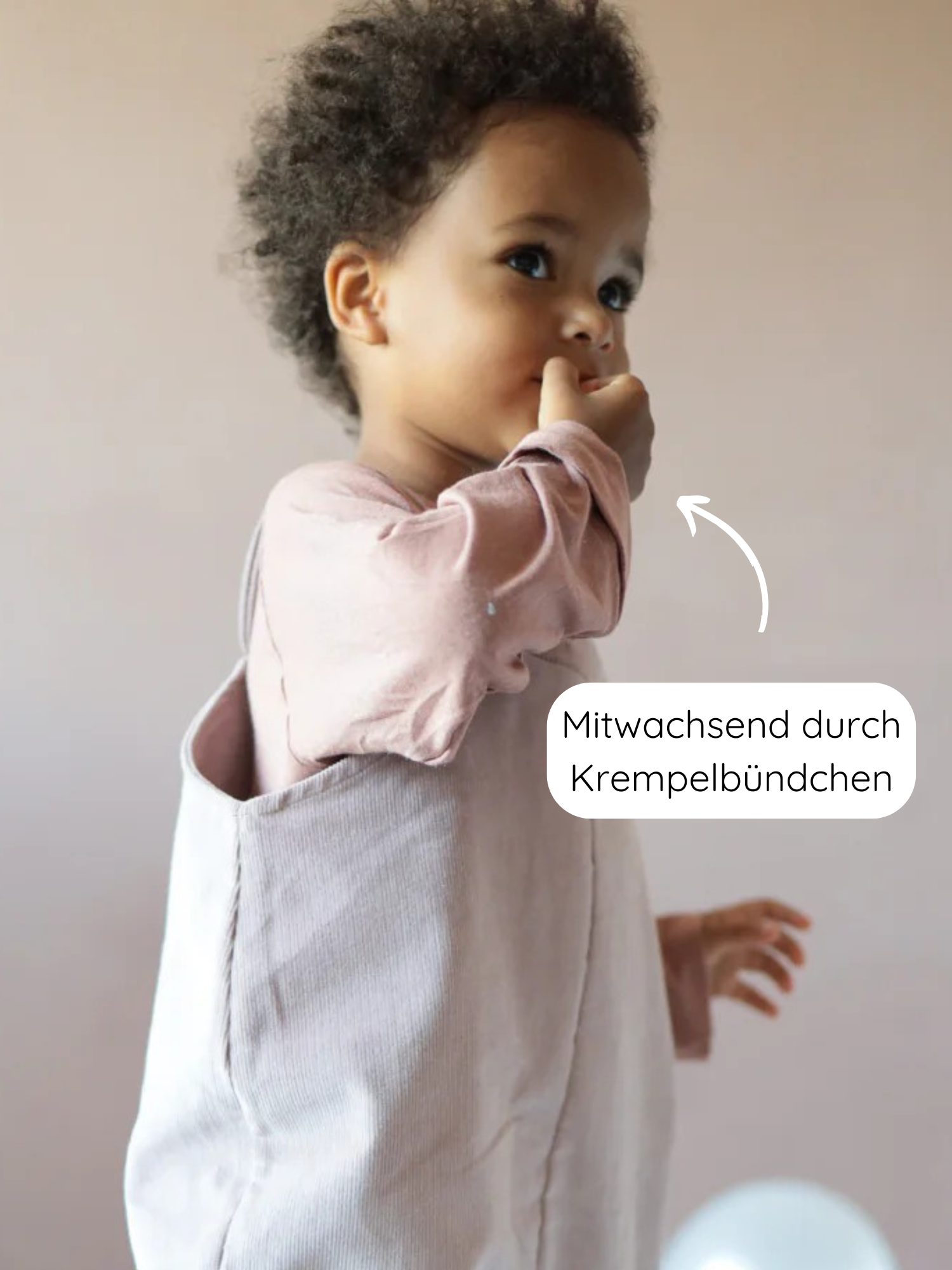Kleinkind im Profil trägt das Wickelshirt aus Kaschmir-Mix - Zimt mit mitwachsenden krempelbaren Bündchen, über einem langärmeligen Shirt. Ein Pfeil zeigt auf die verstellbaren Ärmelbündchen.