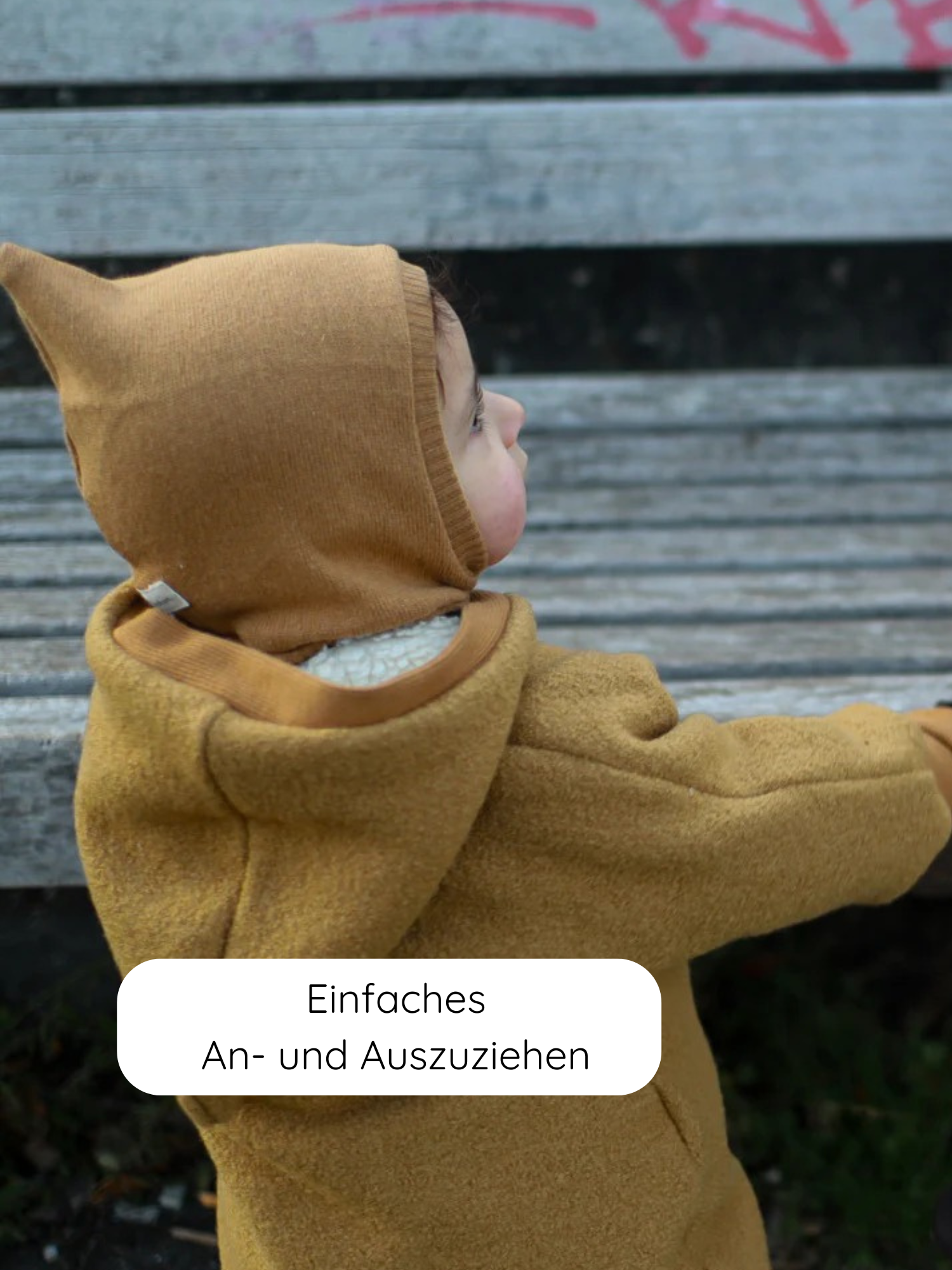 Kleinkind im Profil auf einer Bank, trägt den Winter Wollwalk Overall mit Teddyfutter - Honig, erkennbar an der spitzen Kapuze. Text: „Einfaches An- und Ausziehen“.