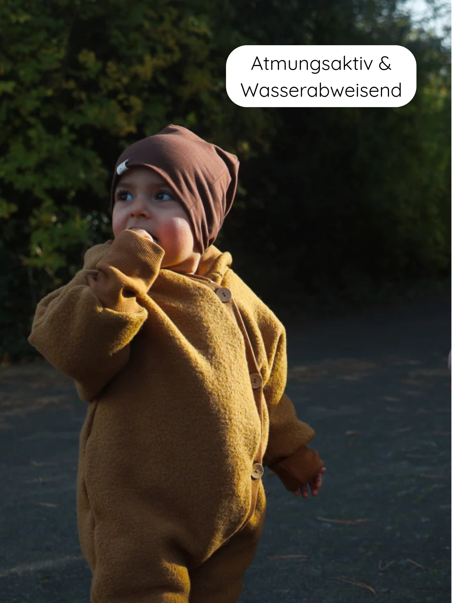 Kleinkind im Winter Wollwalk Overall mit Teddyfutter - Honig mit Holzknöpfen und breiten Bündchen, trägt eine braune Strickmütze. Beschriftung: Atmungsaktiv & Wasserabweisend.
