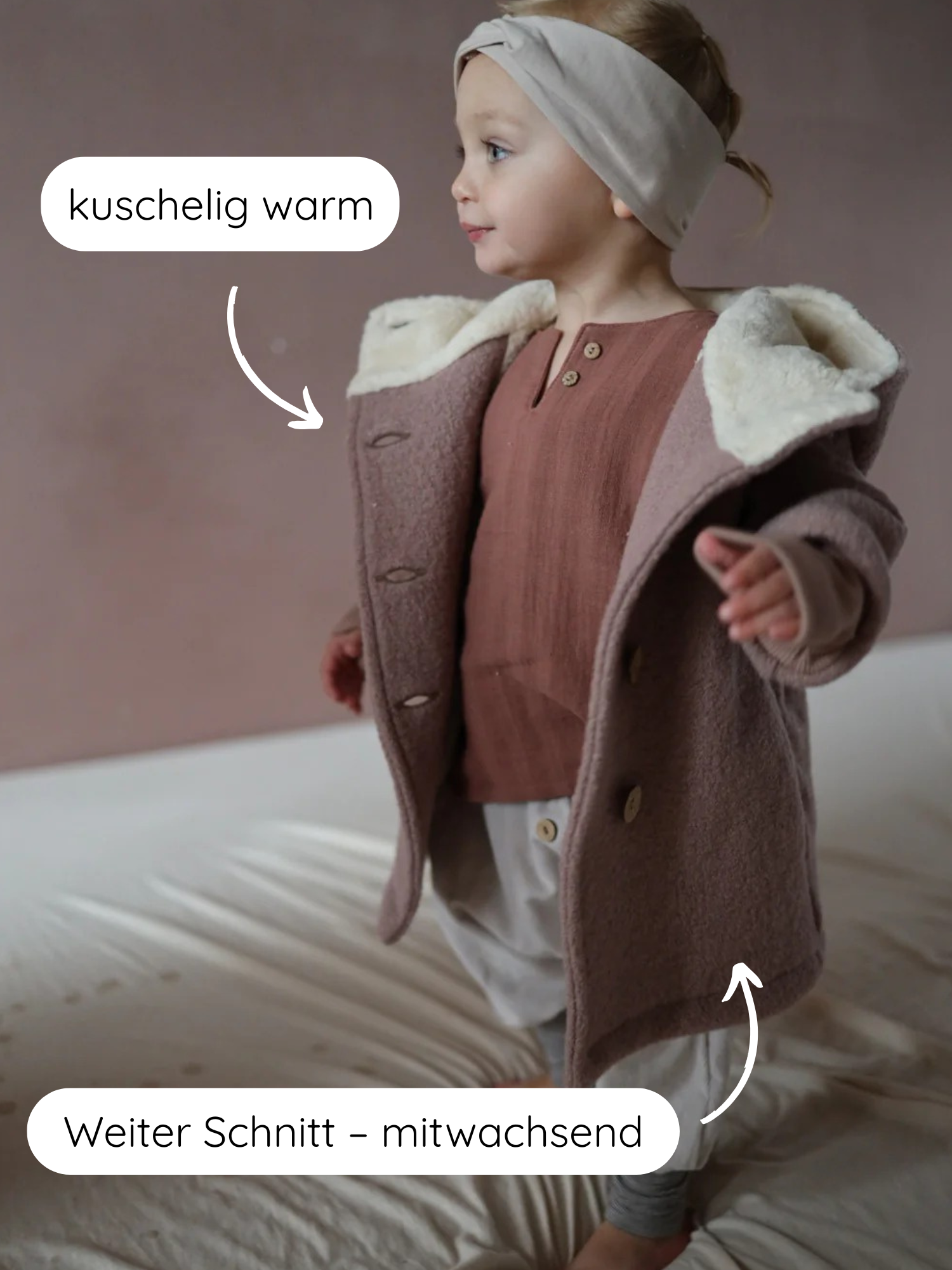 Kleinkind steht auf Bett, trägt Winter Wichteljacke Wollwalk - Puder mit creme-farbenem Teddyfutter, rustfarbenem Shirt, grauer Hose mit Holzknöpfen, grauen Leggings und beigem Stirnband. Jacke ist kuschelig, wetterfest.