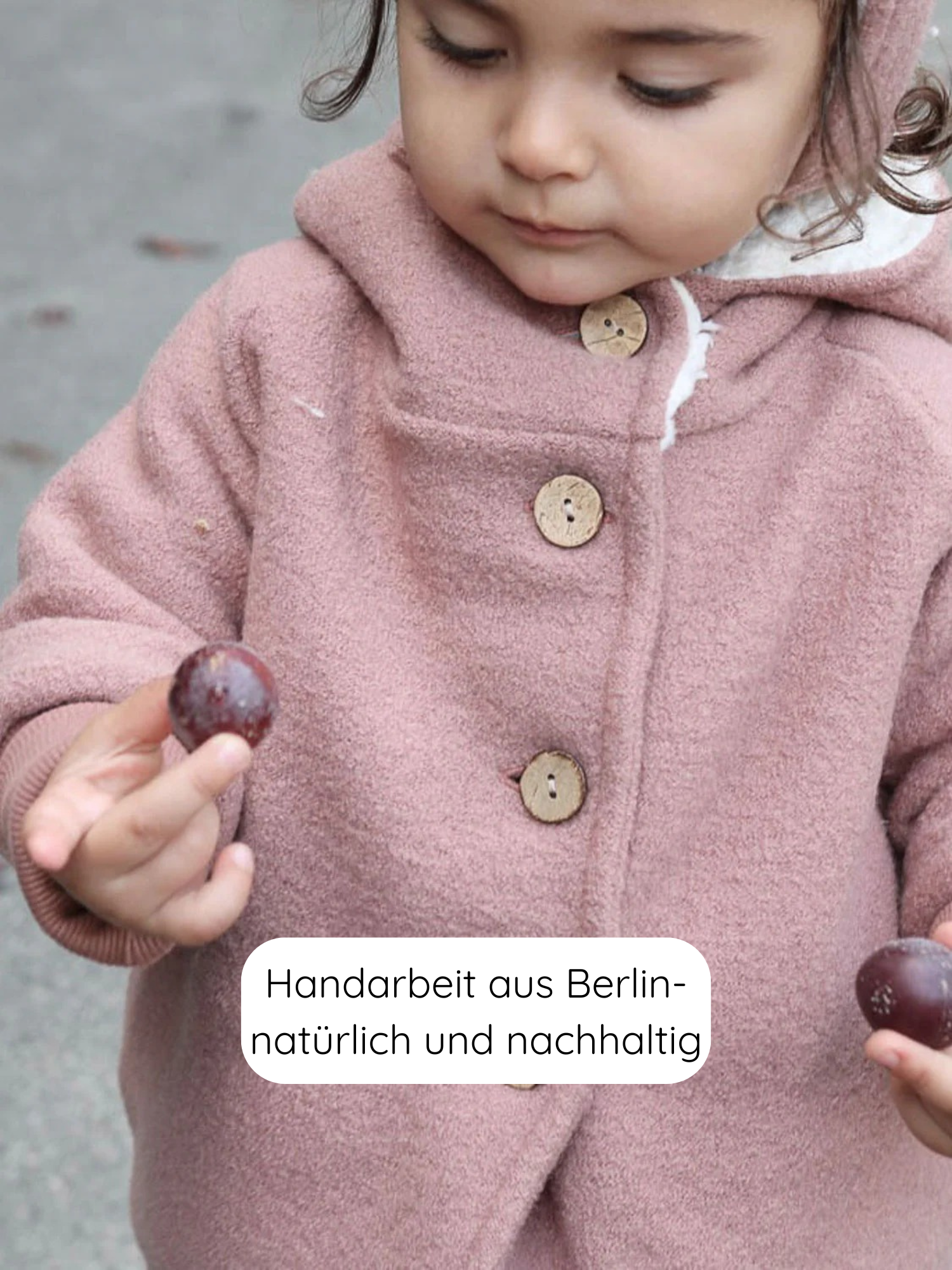 Kleinkind trägt die Winter Wichteljacke Wollwalk - Puder mit hölzernen Knöpfen und spitzer Kapuze, hält zwei kleine lila Früchte. Text: “Handarbeit aus Berlin – natürlich und nachhaltig”.
