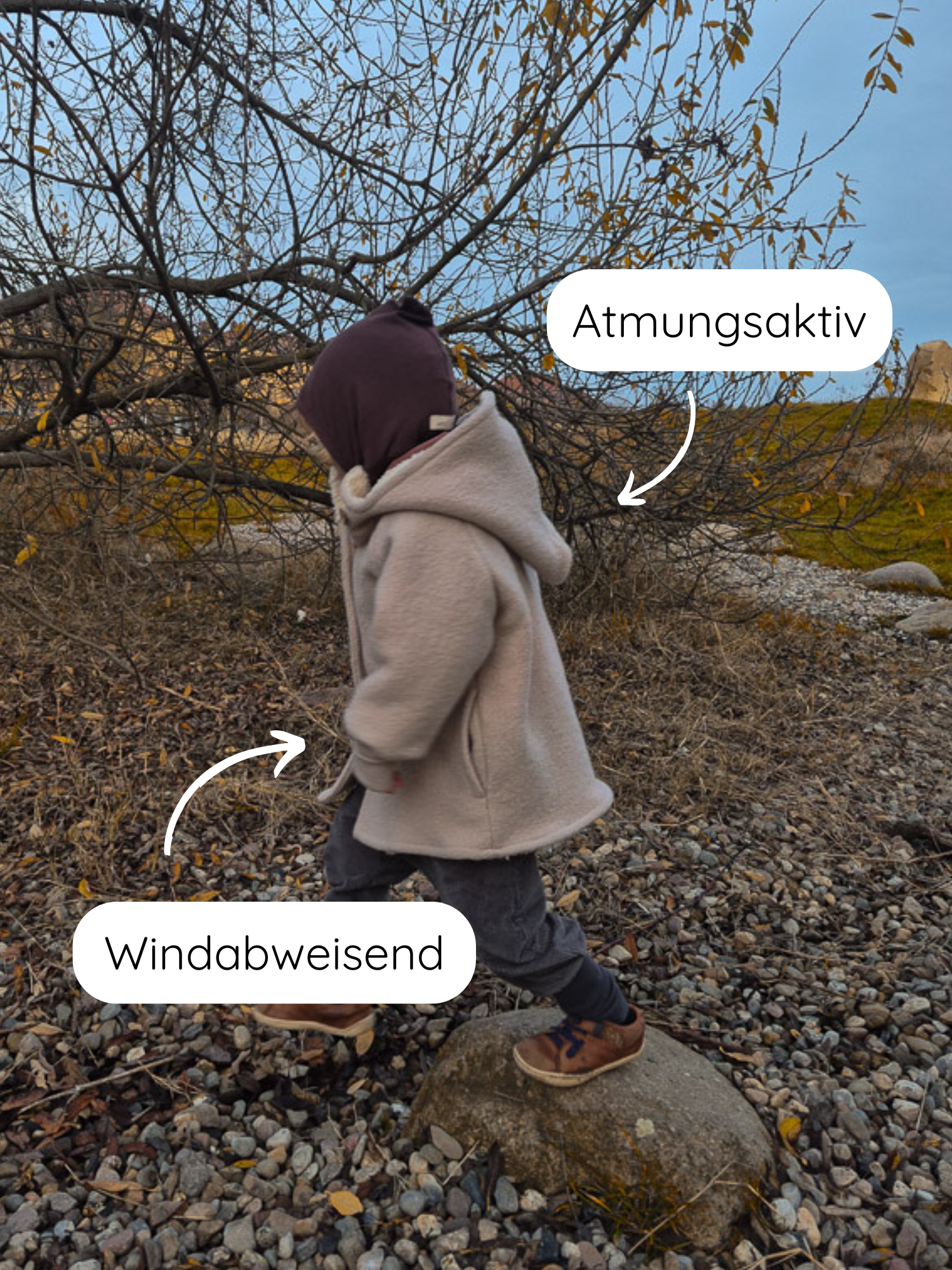 Ein kleines Kind trägt die Winter Wichteljacke Wollwalk - Sand mit Kapuze, atmungsaktiv und windabweisend, beim Abstieg von einem Felsen auf kiesigen Boden, umgeben von kahlen Ästen.