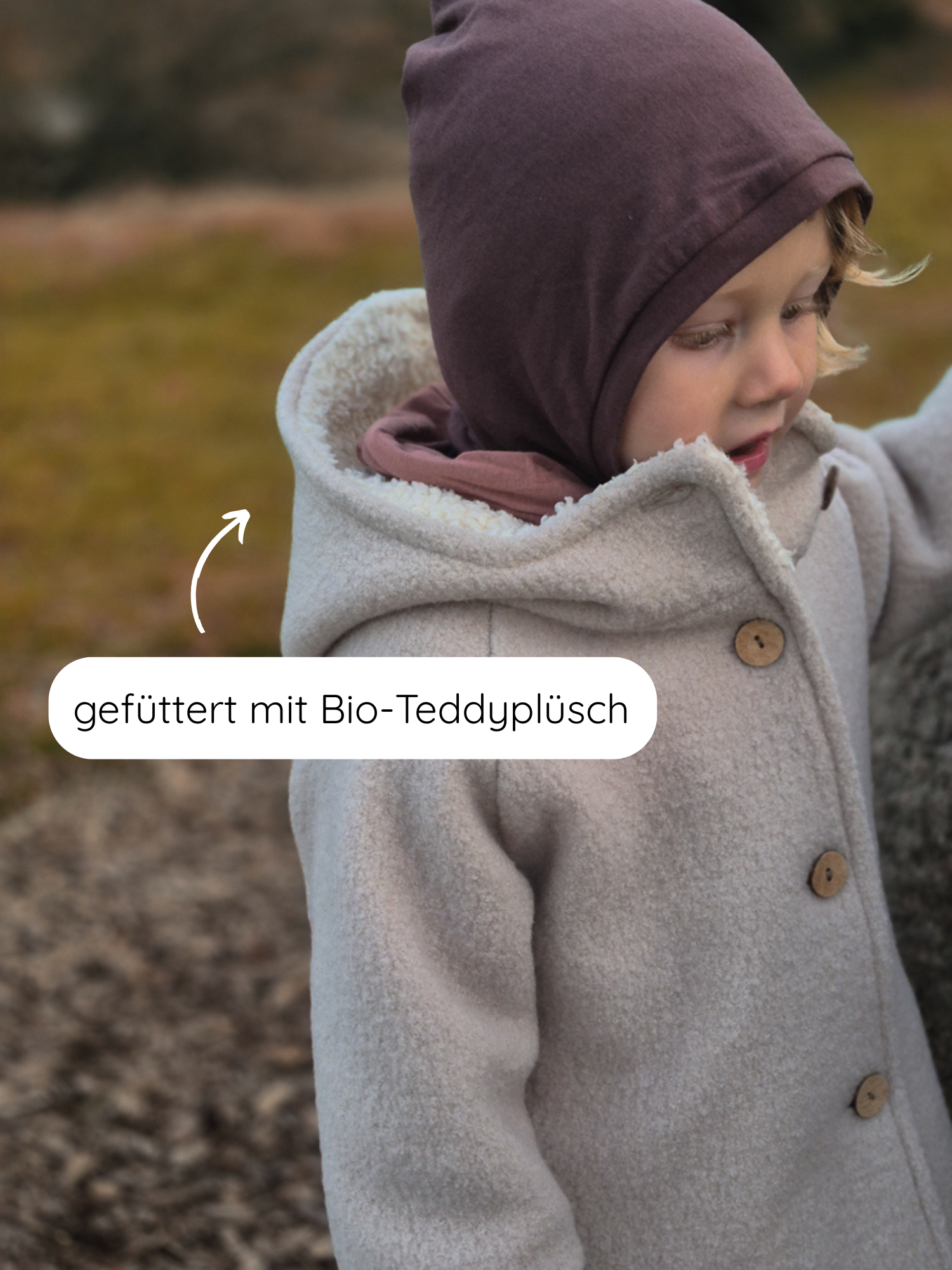 Kleinkind trägt draußen die Winter Wichteljacke Wollwalk - Sand, mit Holzknöpfen und Kapuze, innen mit Bio-Teddyplüsch gefüttert, ideal für kalte Temperaturen. Ein Pfeil zeigt das kuschelige Innenfutter.