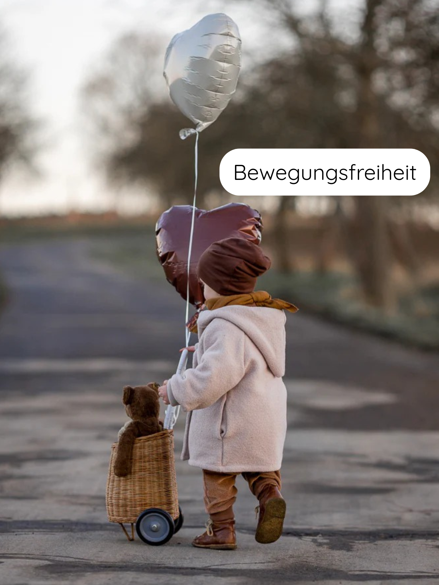 Ein Kind zieht einen Bollerwagen mit Teddy, hält Herzluftballons. Es trägt die Winter Wichteljacke Wollwalk - Sand, warm gefüttert, ideal für kalte Tage, auf einem Landweg.