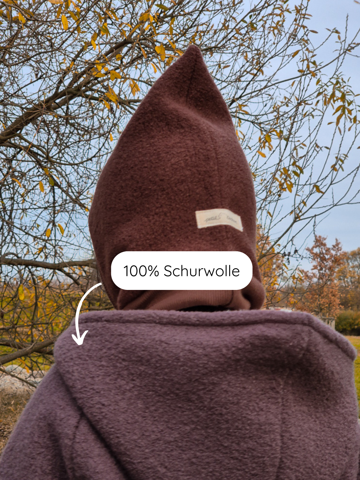 Person von hinten in Herbstlandschaft mit Winter Wichteljacke Wollwalk mit Teddyfutter - Malve, spitzer Kapuze und 100 % Schurwolle, betont durch eingeblendeten Hinweis auf das Material.