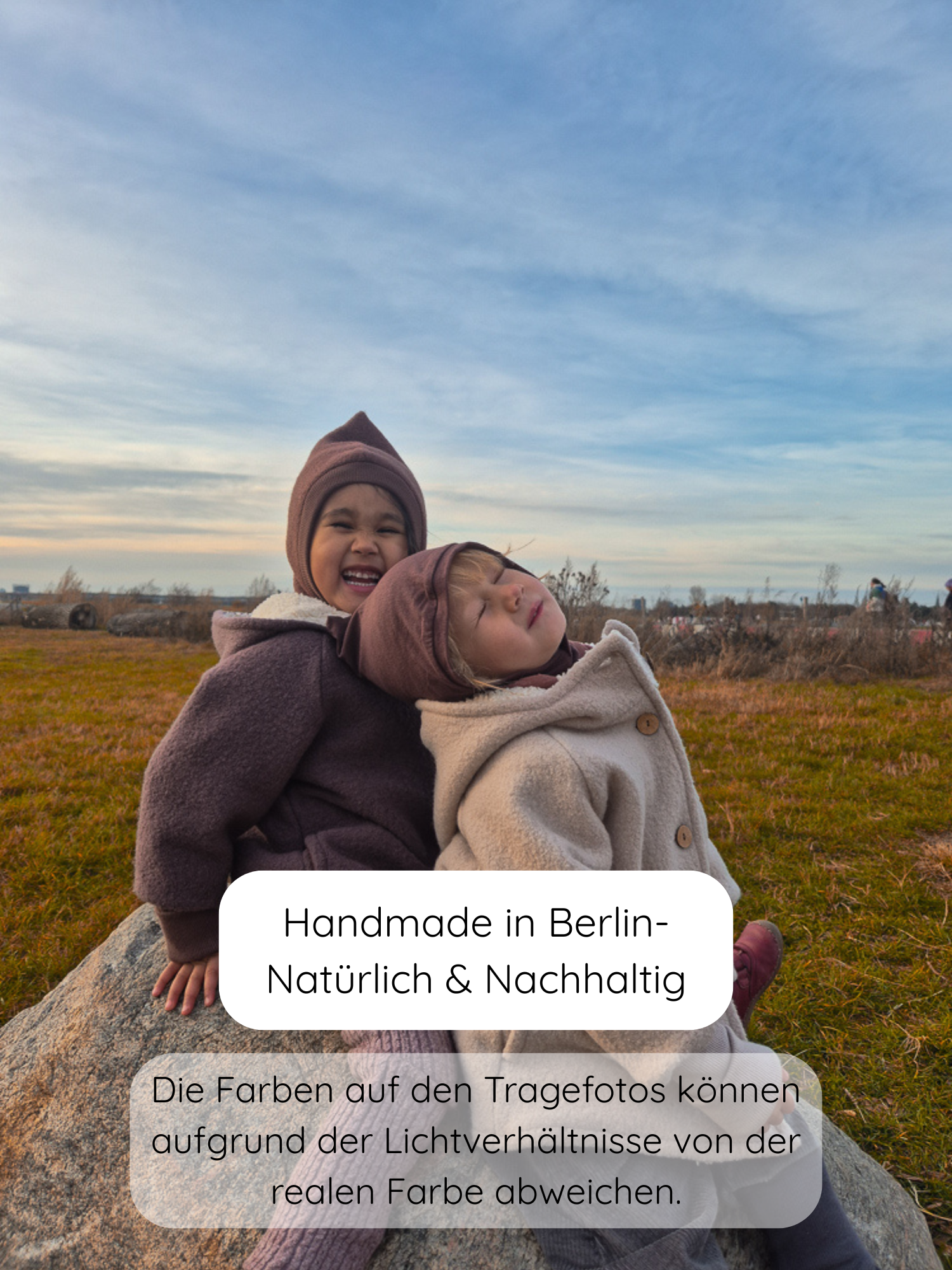 Zwei Kinder in gemütlichen, handgefertigten Winter Wichteljacken Wollwalk mit Teddyfutter - Malve sitzen auf einem Stein im Feld. Die Jacken haben spitze Kapuzen und breite Bündchen.