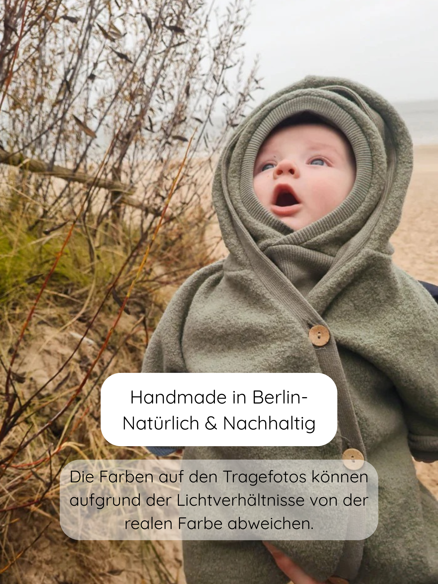 Baby in einem Kuschelanzug Wollwalk - Pistazie aus 100% Schurwolle mit Kapuze, sitzt auf einem Strand oder Düne. Der Overall ist großzügig geschnitten und hat krempelbare Bündchen.
