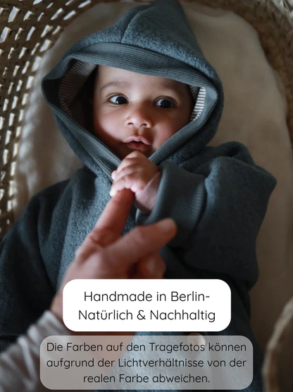 Kuschelanzug Wollwalk - Taube: Baby in einem gewebten Moseskorb, trägt einen weichen, schiefergrauen Kapuzenanzug und hält einen Erwachsenenfinger fest. Text: Handmade in Berlin – Natürlich & Nachhaltig.