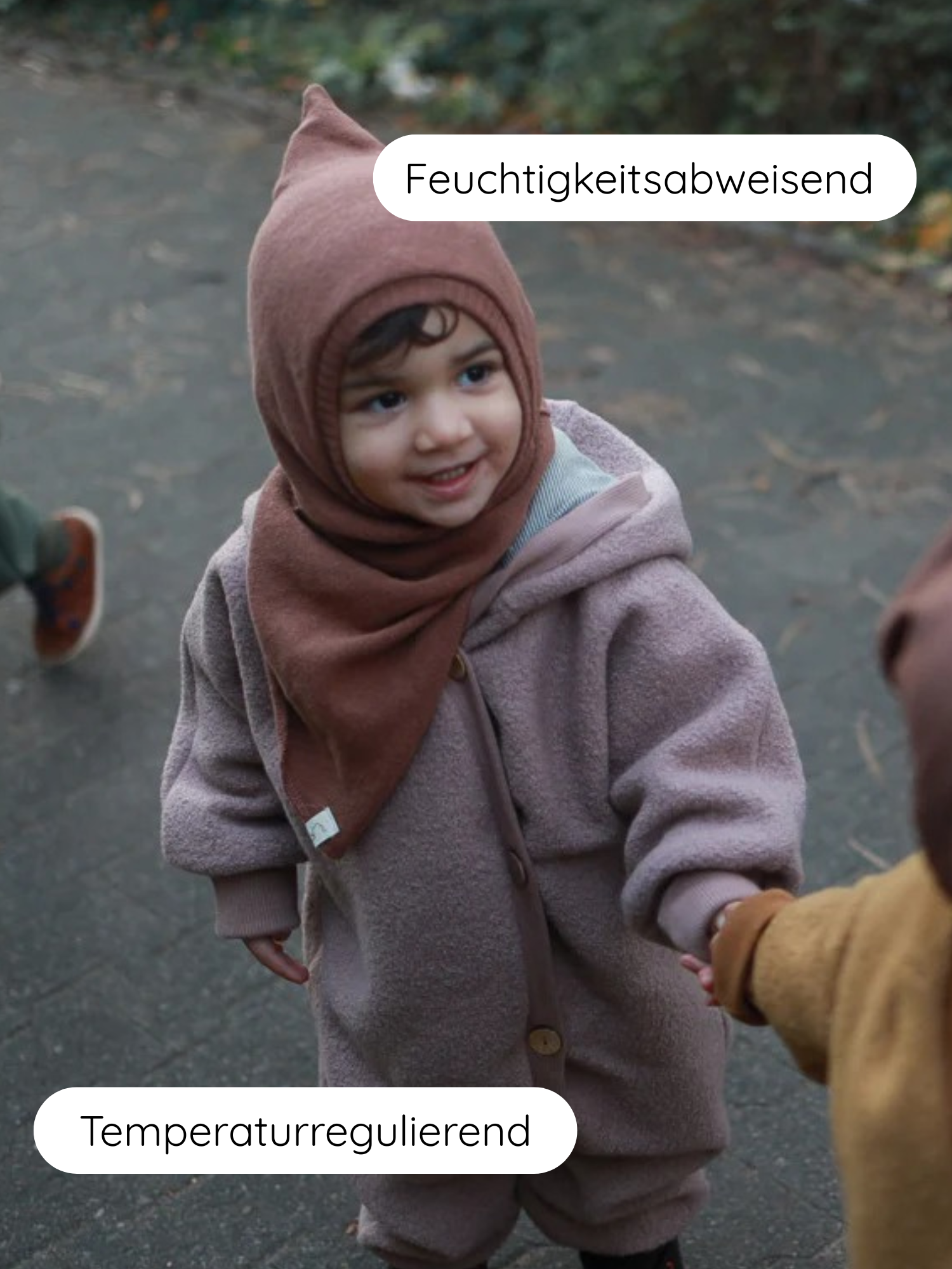 Kleinkind im Kuschelanzug Wollwalk - Puder mit spitzer Kapuze, lächelnd und Hand haltend. Der Anzug ist feuchtigkeitsabweisend und temperaturregulierend, ideal für kühle Tage im Freien.