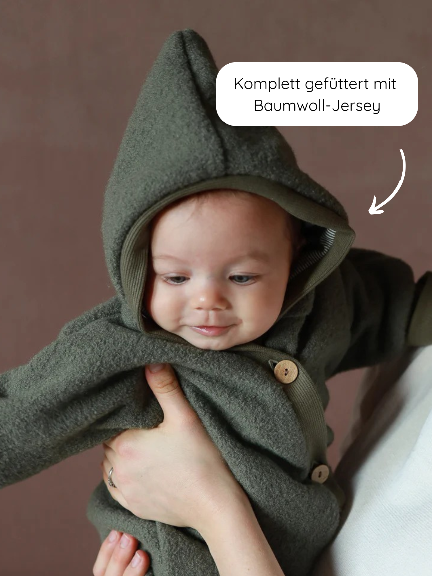 Kuschelanzug Wollwalk - Pistazie: Baby in einem mossgrünen Fleece-Einteiler mit spitzer Kapuze, Holzknöpfen vorn; komplett gefüttert mit Baumwoll-Jersey. Gehalten von Erwachsenenhand, vor neutralem Hintergrund.