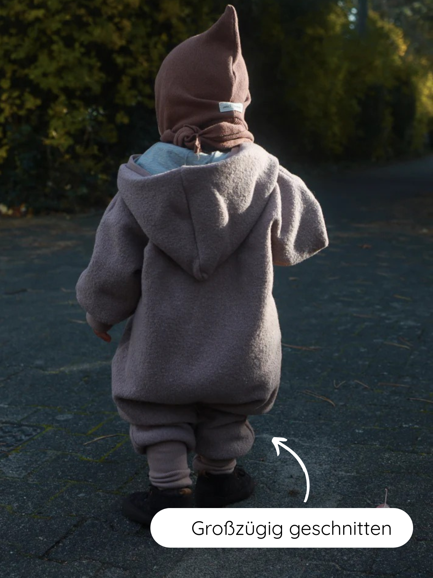 Ein Kleinkind steht auf einem gepflasterten Weg, trägt den Kuschelanzug Wollwalk - Puder, einen locker sitzenden Overall aus Schurwolle mit Kapuze und spitzem Mütze, großzügig geschnitten für Bewegungsfreiheit.