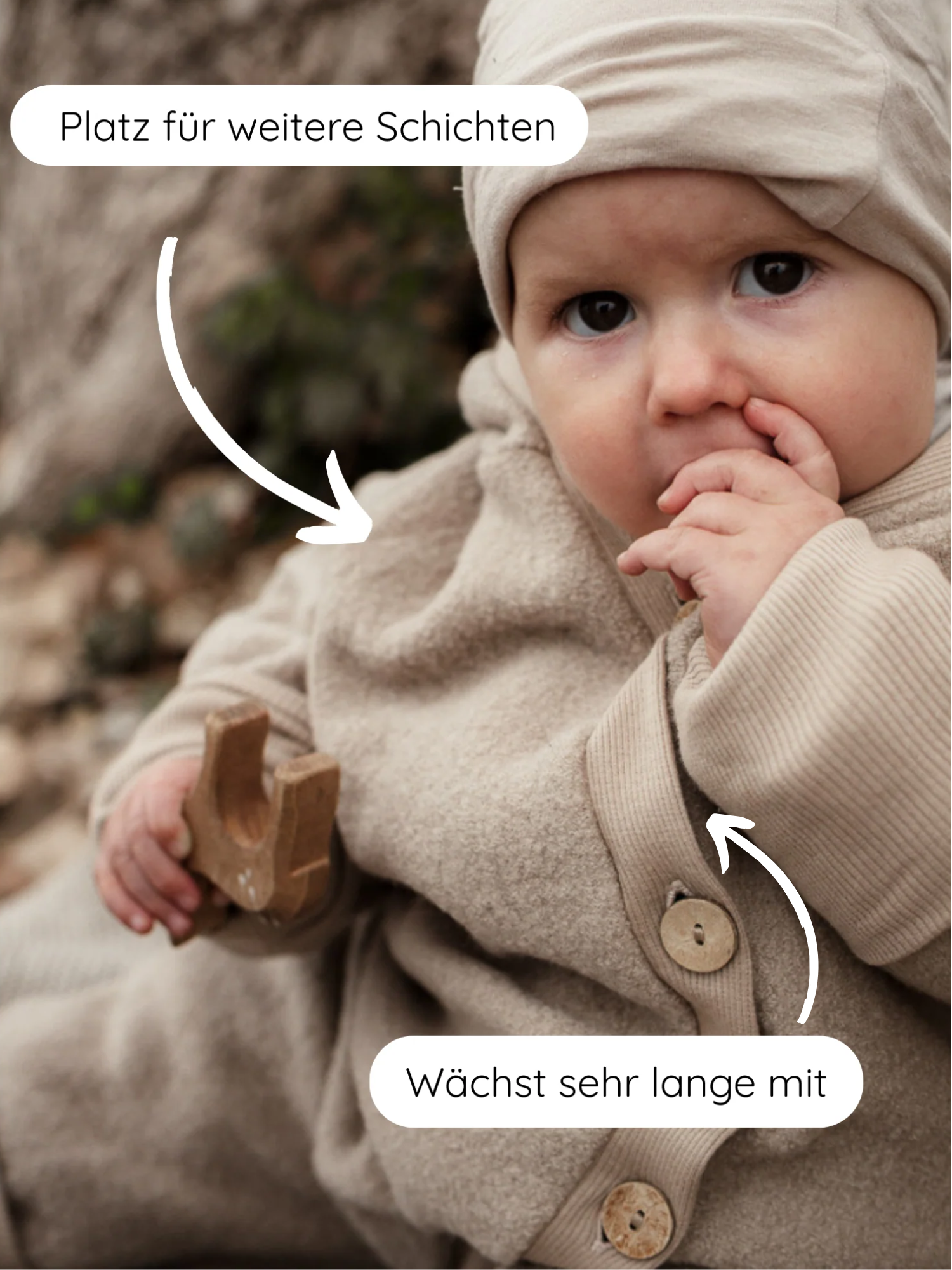 Kleinkind im Kuschelanzug Wollwalk - Sand, sitzt im Freien, trägt einen beigen, gerippten Einteiler mit passender Mütze, hält ein Holzspielzeug, weiße Pfeile betonen Platz für Schichten und mitwachsende Bündchen.