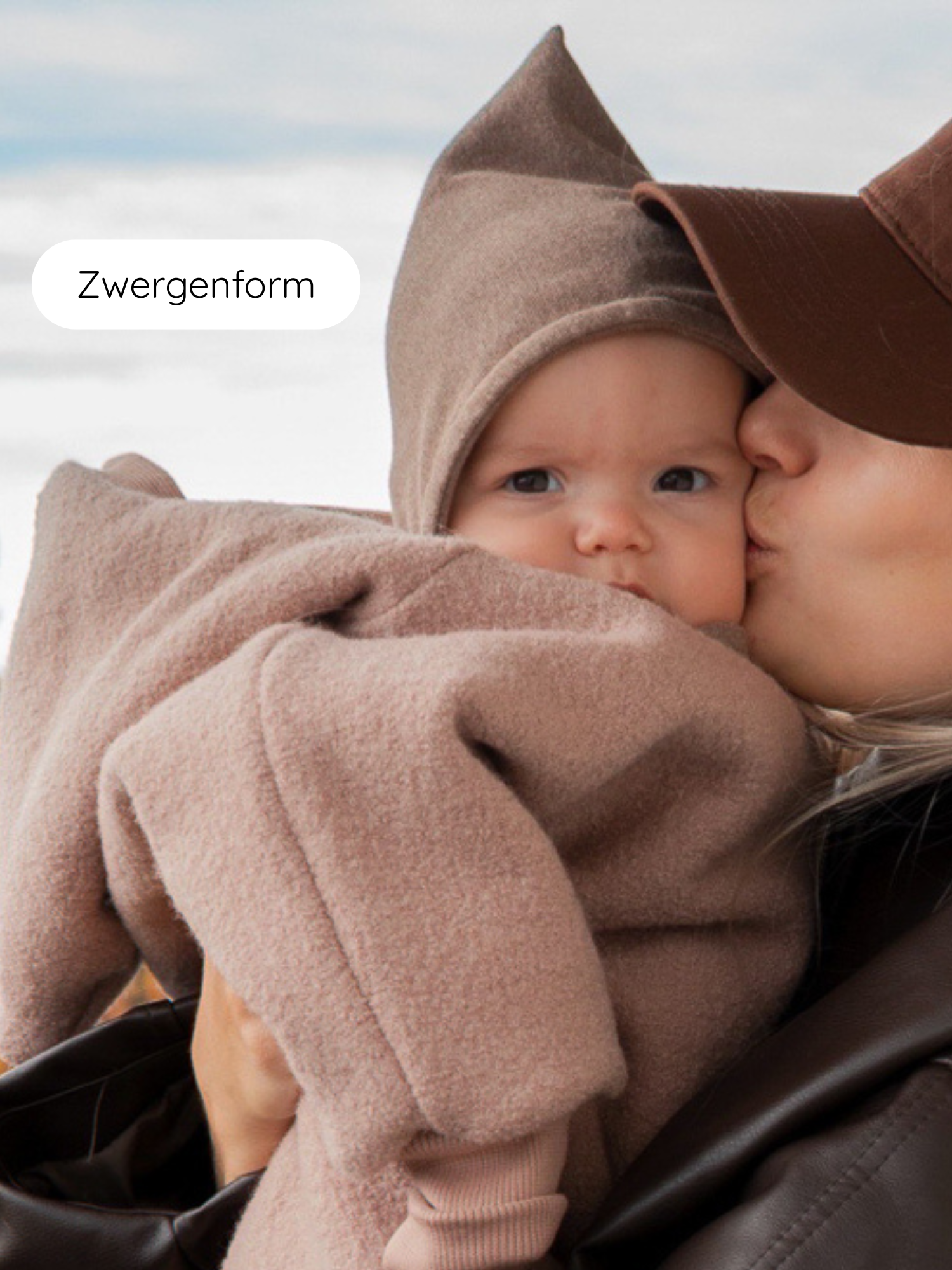 Eine Mutter trägt ihr Baby in einem weichen, beigen Wrap. Das Baby trägt eine Zwergenmütze Feinstrick - Hafer Meliert, die Kopf und Ohren warm hält.