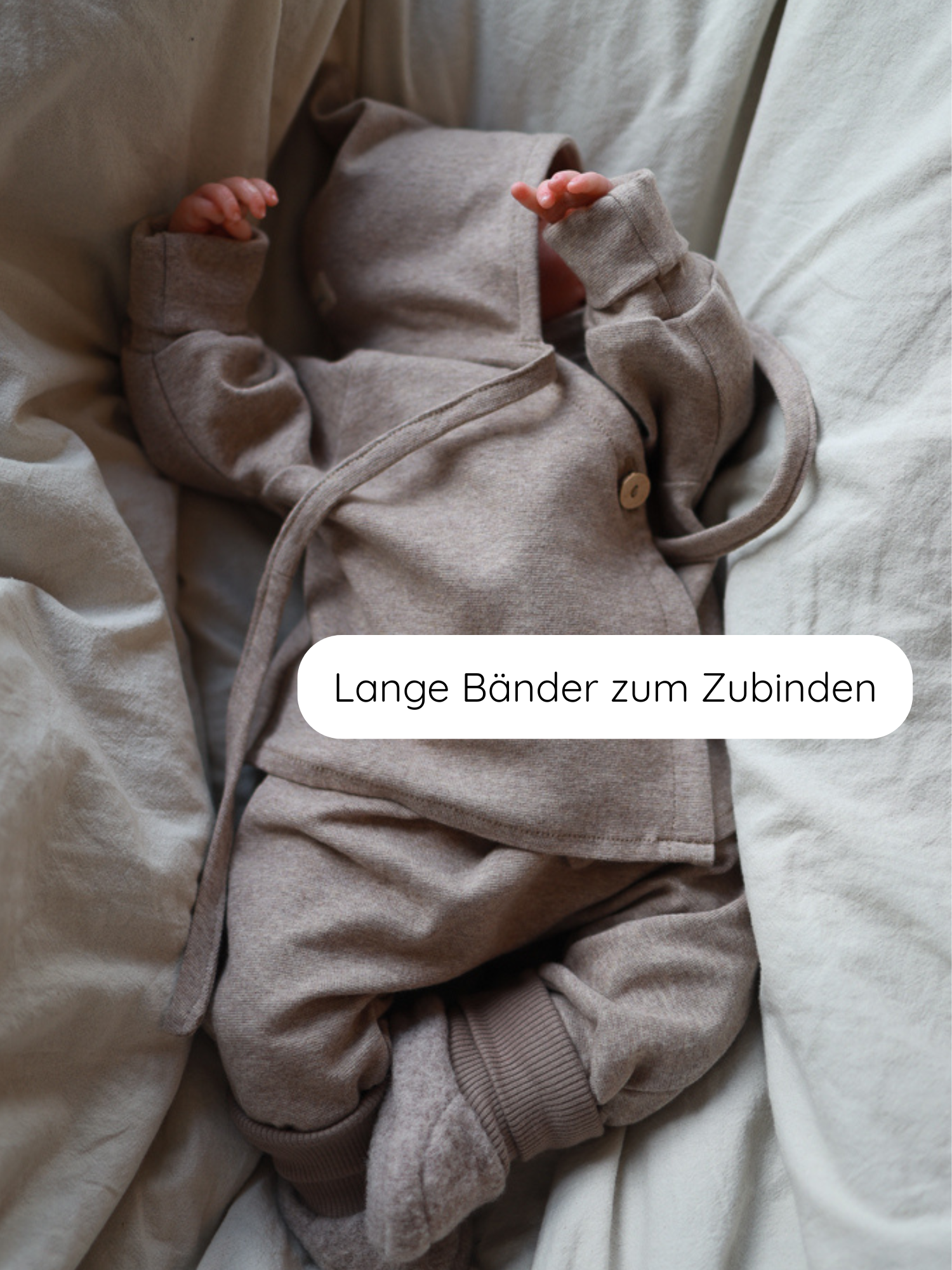 Zwergenmütze Feinstrick - Hafer Meliert: Ein Baby in neutral-beigem Strickoutfit mit Wickelkapuze, Joggerhosen und passenden Booties. Die Mütze schützt Kopf und Ohren, gefertigt aus weichem Feinstrick-Jersey.