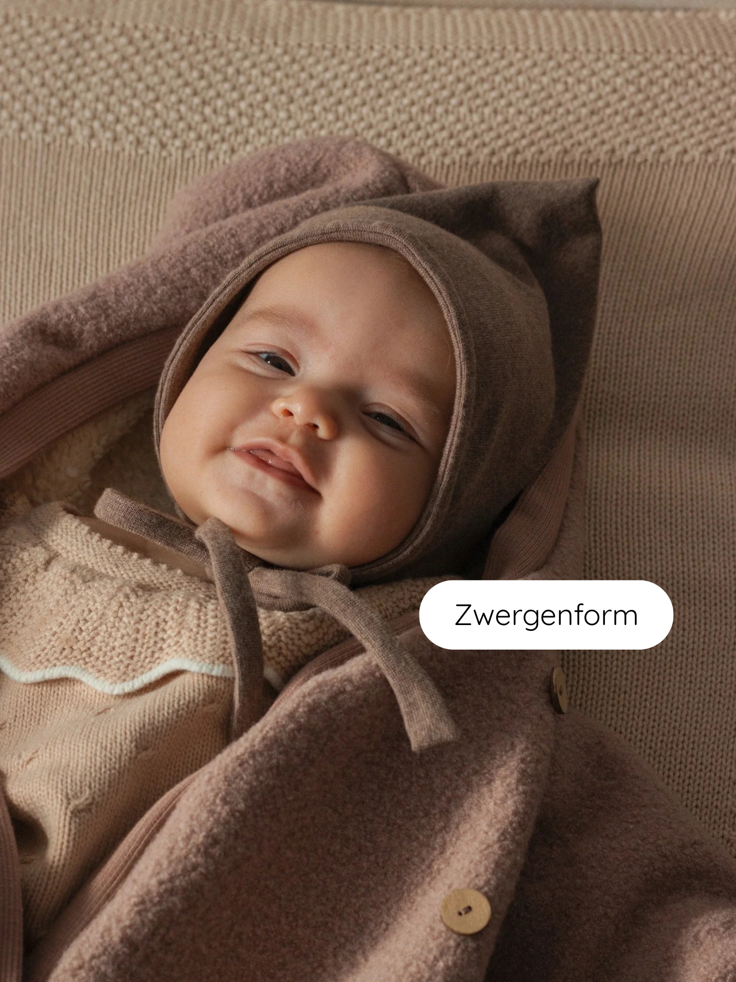 Ein lächelndes Baby trägt die Zwergenmütze Feinstrick - Praline Meliert, aus weichem Jersey, mit charakteristischem Zipfel, liegend auf einer beigen, strukturierten Unterlage.