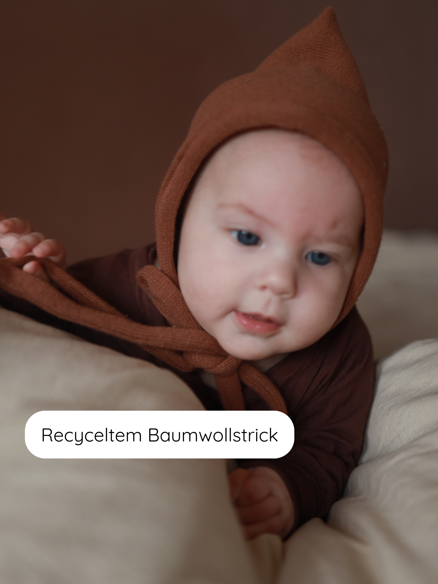 Kleines Baby in Nougatfarbenem Zwergenmütze Strick aus recycelter Baumwolle, liegend auf weicher Oberfläche. Mütze mit elastischem Stoff und Bindeband für perfekte Passform, ideal für Herbst und Winter.