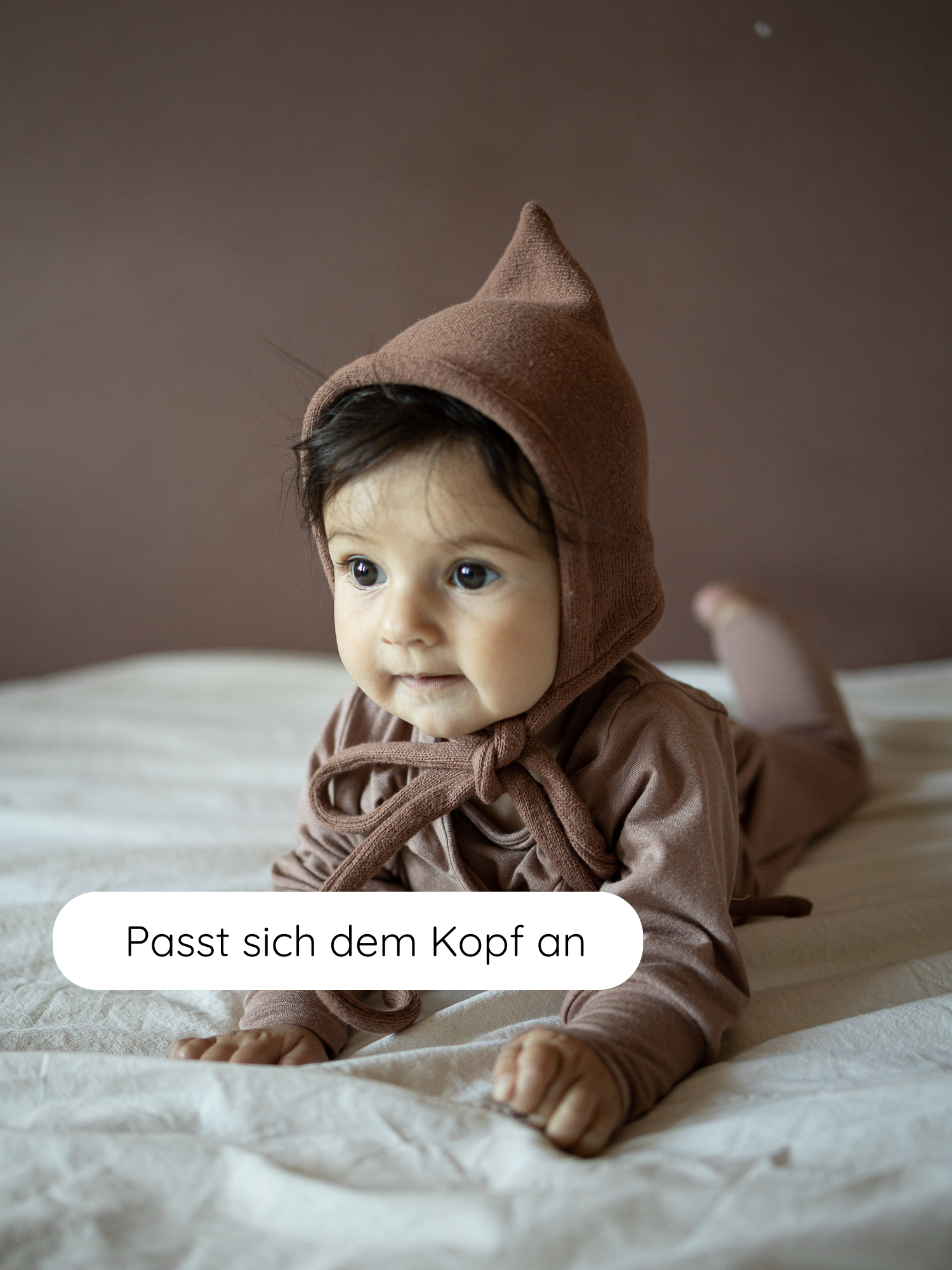 Ein Baby liegt bäuchlings auf einer weißen Decke, trägt die Zwergenmütze Strick - Nougat, eine weiche, spitz zulaufende Babymütze mit Bindeband, die sich flexibel an den Kopf anpasst.
