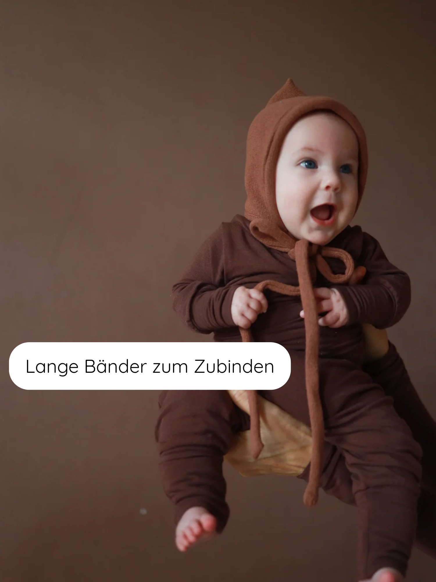 Ein lächelndes Baby trägt die Zwergenmütze Strick - Nougat mit langen Bändern. Die Mütze besteht aus recyceltem Baumwollstrick, ist elastisch und bietet einen gemütlichen Sitz.