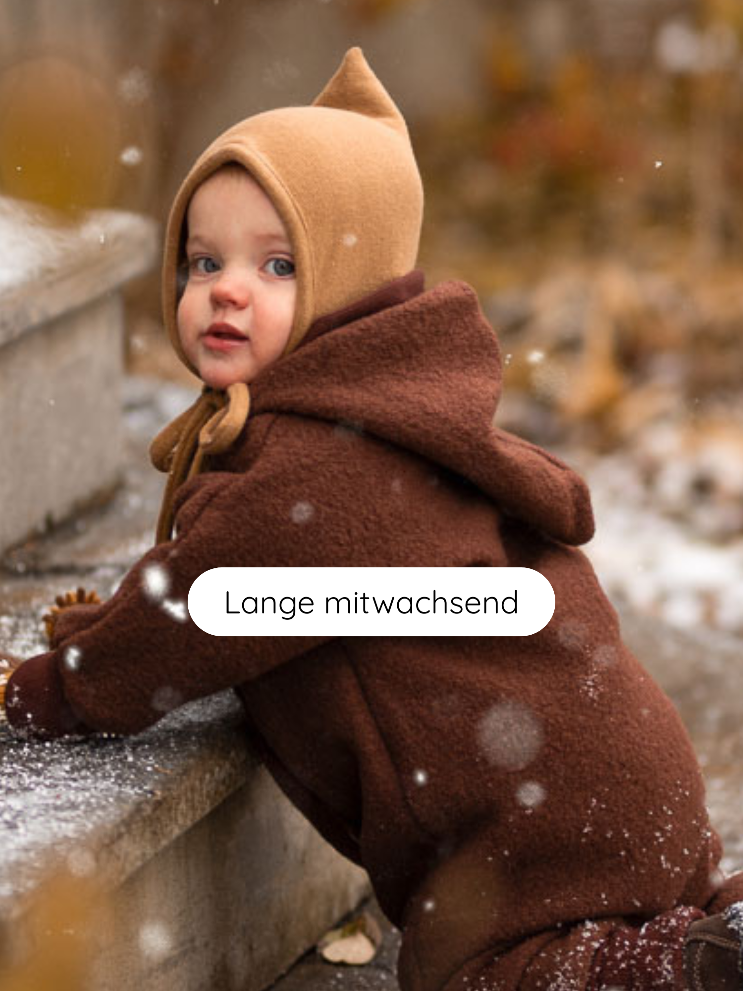 Kleinkind im Zwergenmütze Strick - Sahara, warm eingepackt, lehnt an Stufen im Schnee. Die gestrickte Mütze aus recycelter Baumwolle passt sich dank Bindeband an, ideal für Herbst und Winter.