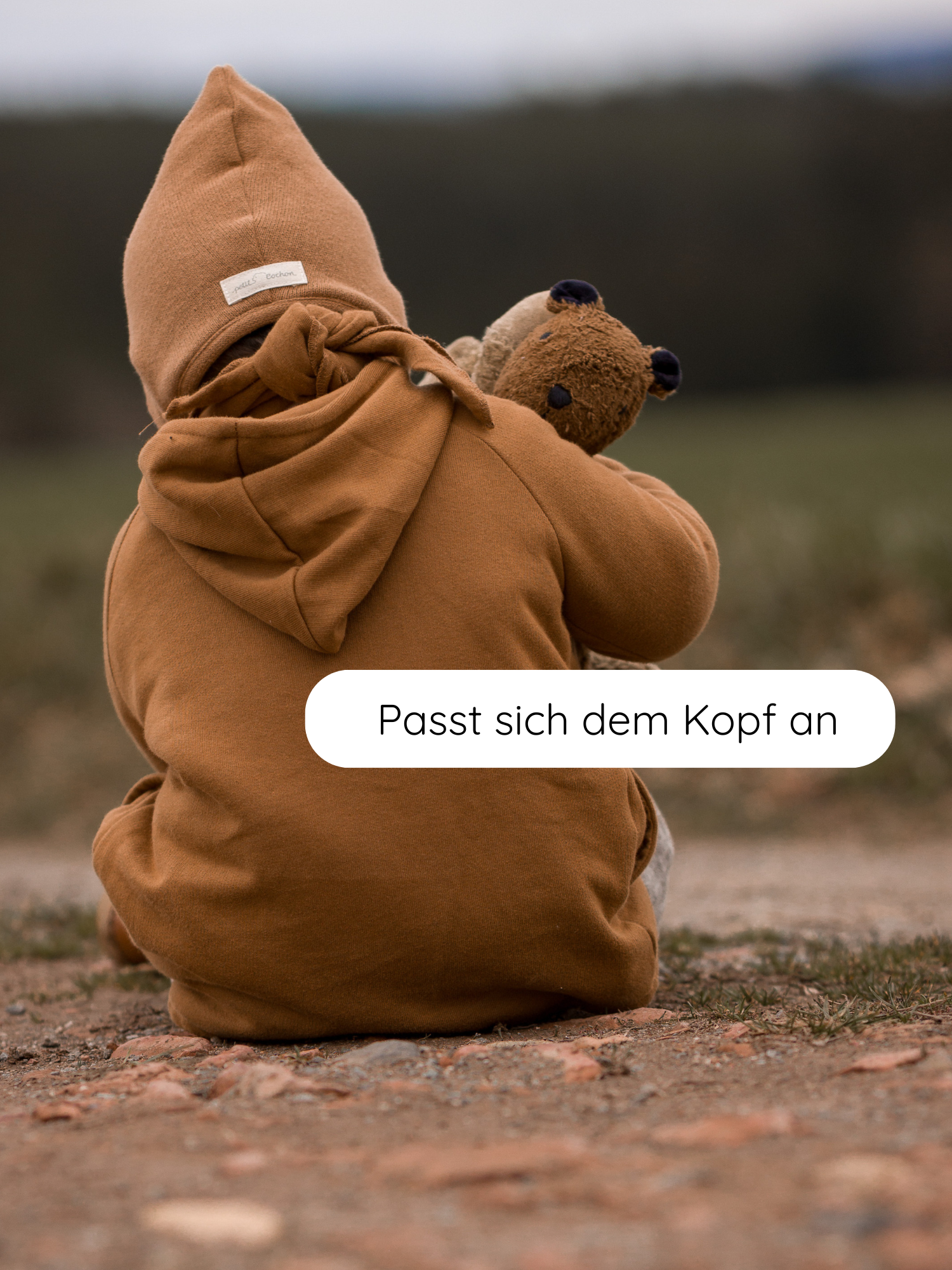Ein Kind von hinten sitzt in einer Zwergenmütze Strick - Sahara, aus recycelter Baumwolle, mit spitzer Kapuze und hält einen Teddy. Sprechblase: “Passt sich dem Kopf an”.