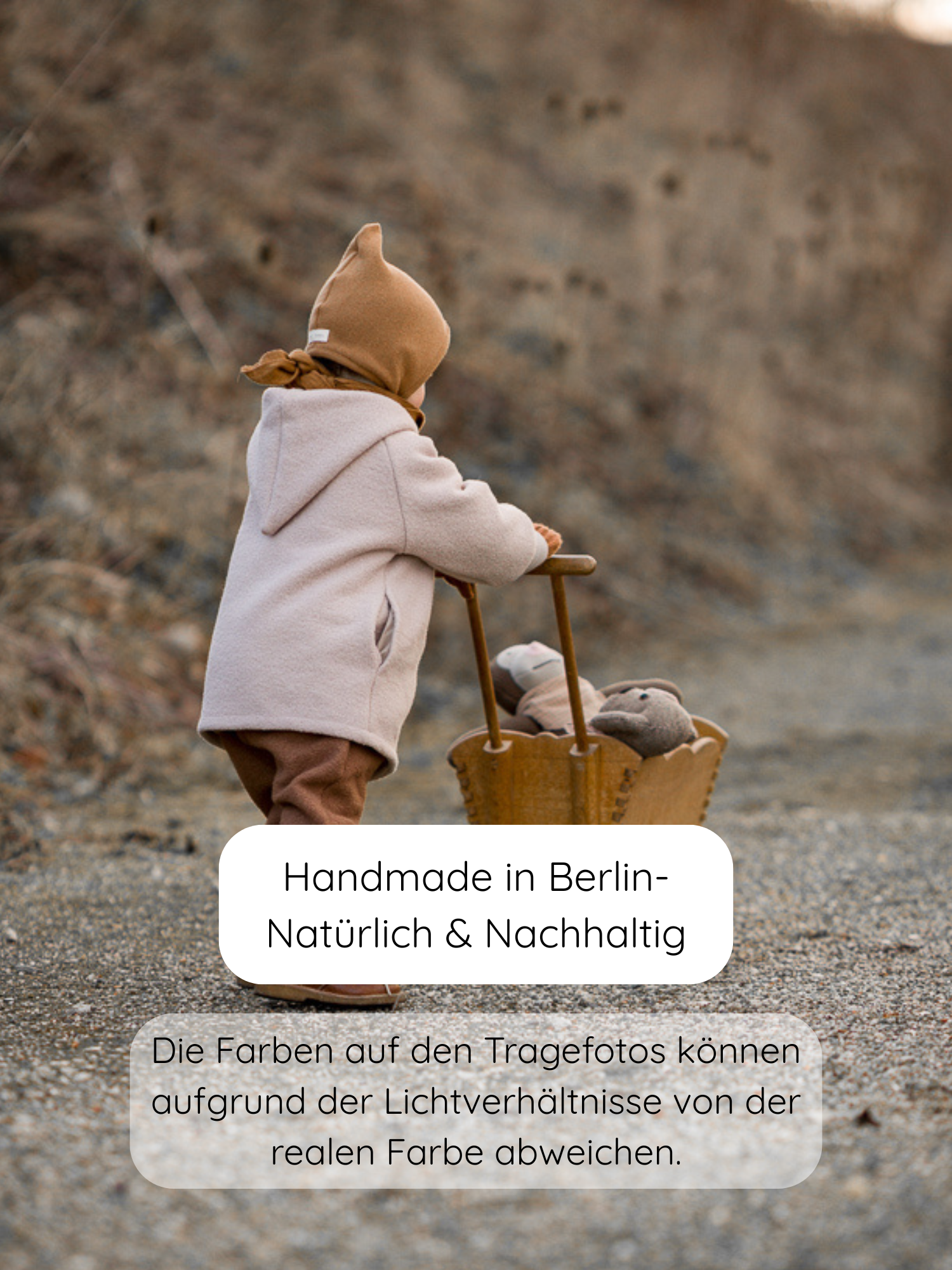 Kleinkind von hinten mit Zwergenmütze Strick - Sahara, stehend auf einem Kiesweg. Es schiebt einen Holzwagen mit Teddybär. Die Mütze ist aus recycelter Baumwolle, perfekt für Herbst und Winter.