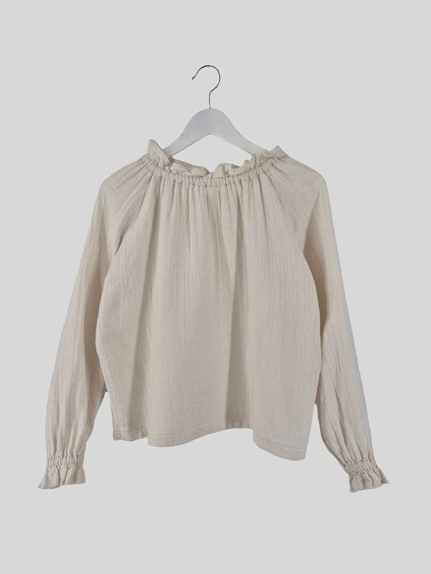 Bluse Bio Musselin Natur: Langärmlige, hellbeige Bluse mit elastischem Kragen und Bündchen, an einem weißen Bügel hängend. Sichtbare Verfärbungen am Kragen, kleiner Fleck und gestopftes Loch auf der Rückseite. Zustand: gut.