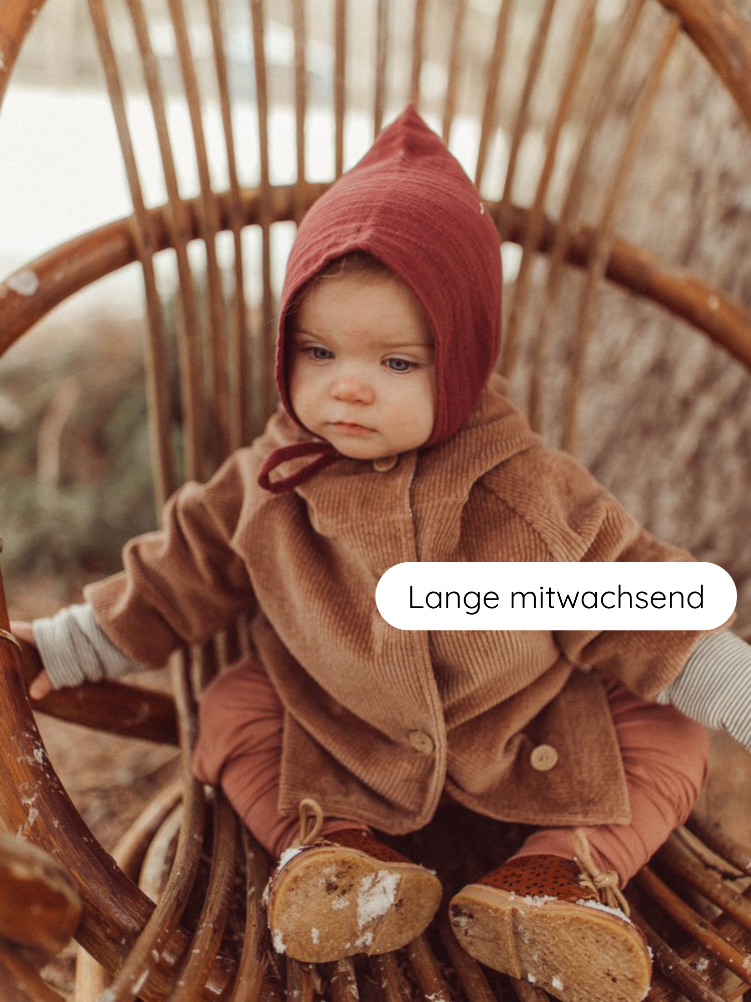 Kleinkind in rustikalem Rattanstuhl trägt Babyknicker Kaschmir-Mix - Zimt mit Klappbündchen, kombiniert mit rustrotem Bonnet, brauner Jacke und Lederschuhen mit Schneestaub.