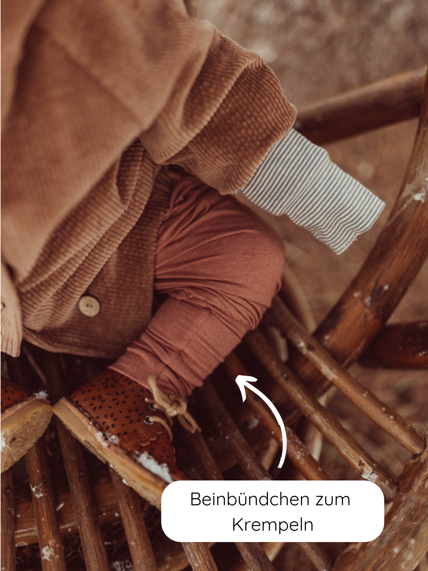 Babyknicker Kaschmir-Mix - Zimt mit umklappbaren Beinbündchen, getragen von einem Kind. Rustfarbene Leggings mit geripptem, aufrollbarem Bündchen, kombiniert mit braunen Lederschuhen, auf einem Holzstuhl sitzend.